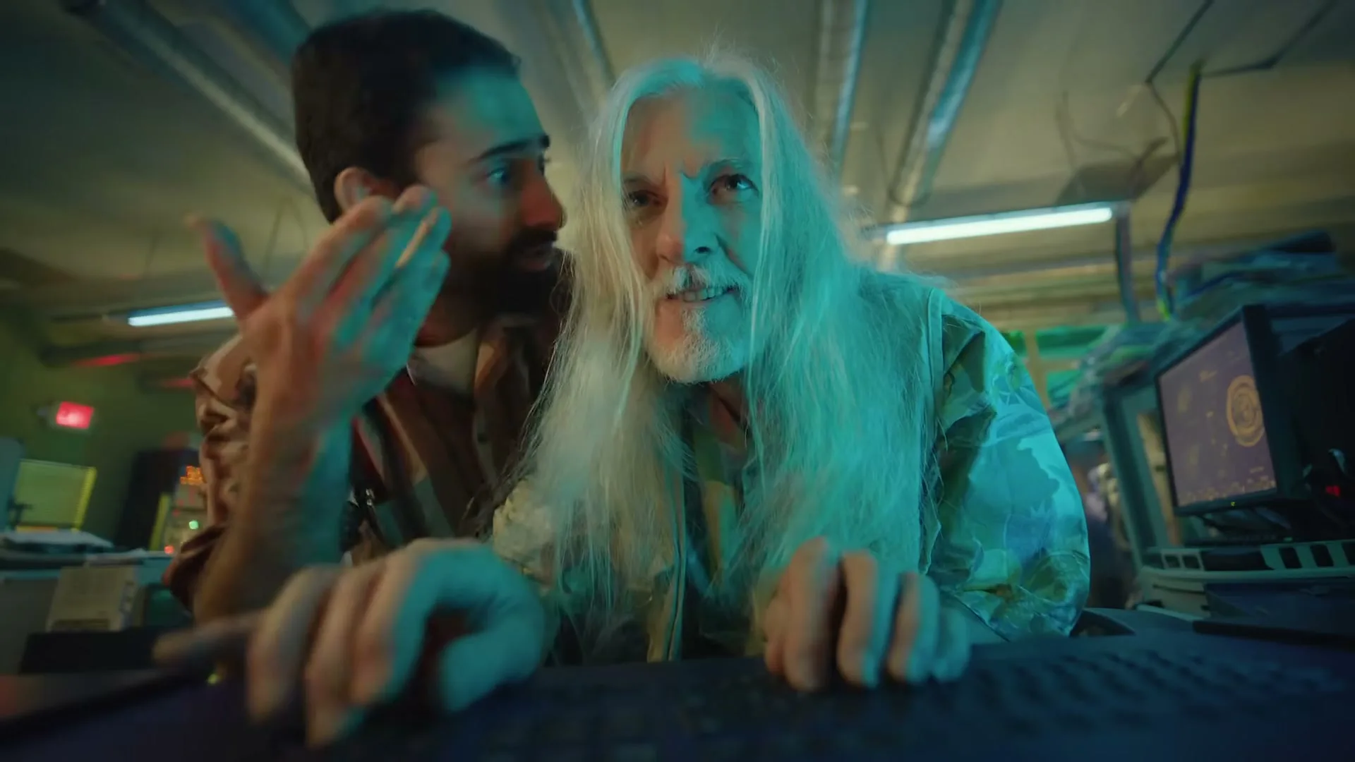 Two men, one with long white hair and one with dark hair and a beard, are looking intently at a computer screen in a dimly lit, cluttered room.
