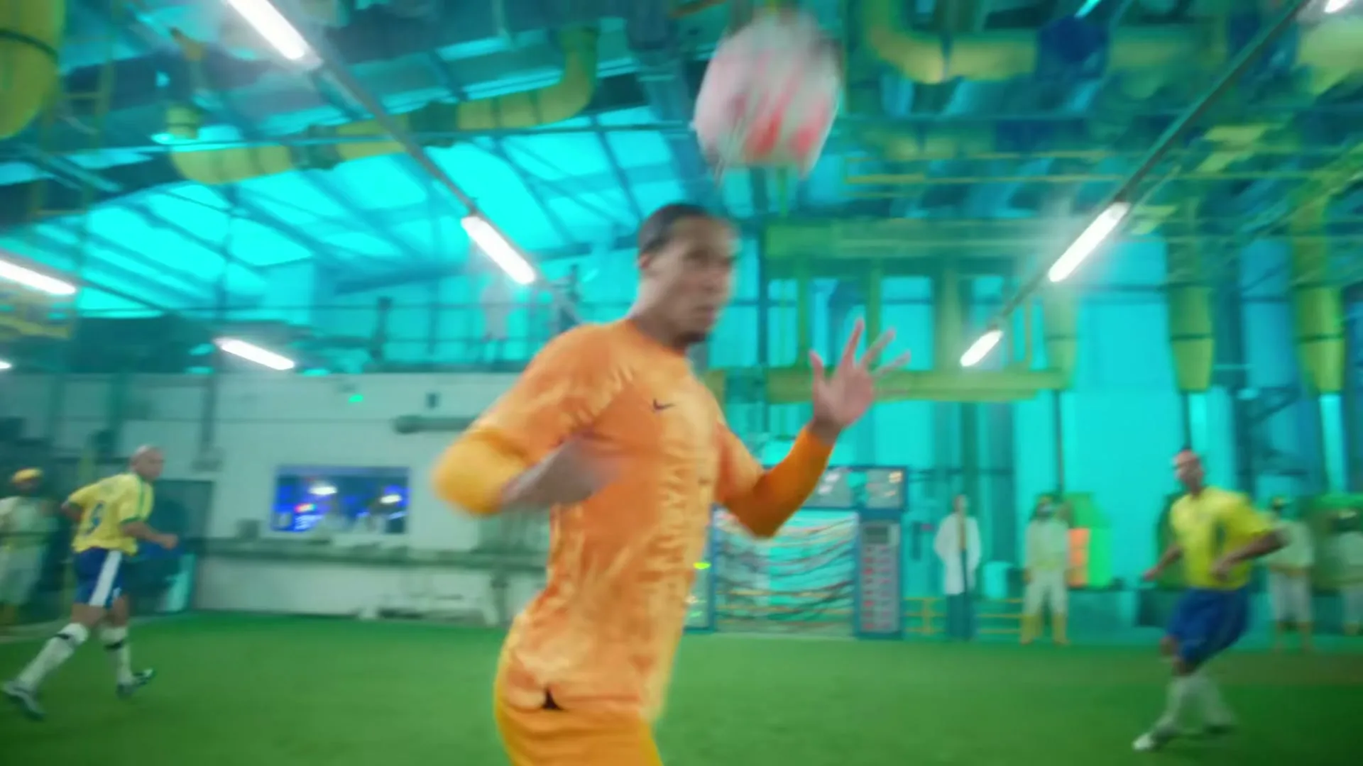 A male soccer player in an orange uniform heads a soccer ball in a brightly lit, futuristic-looking indoor soccer field.