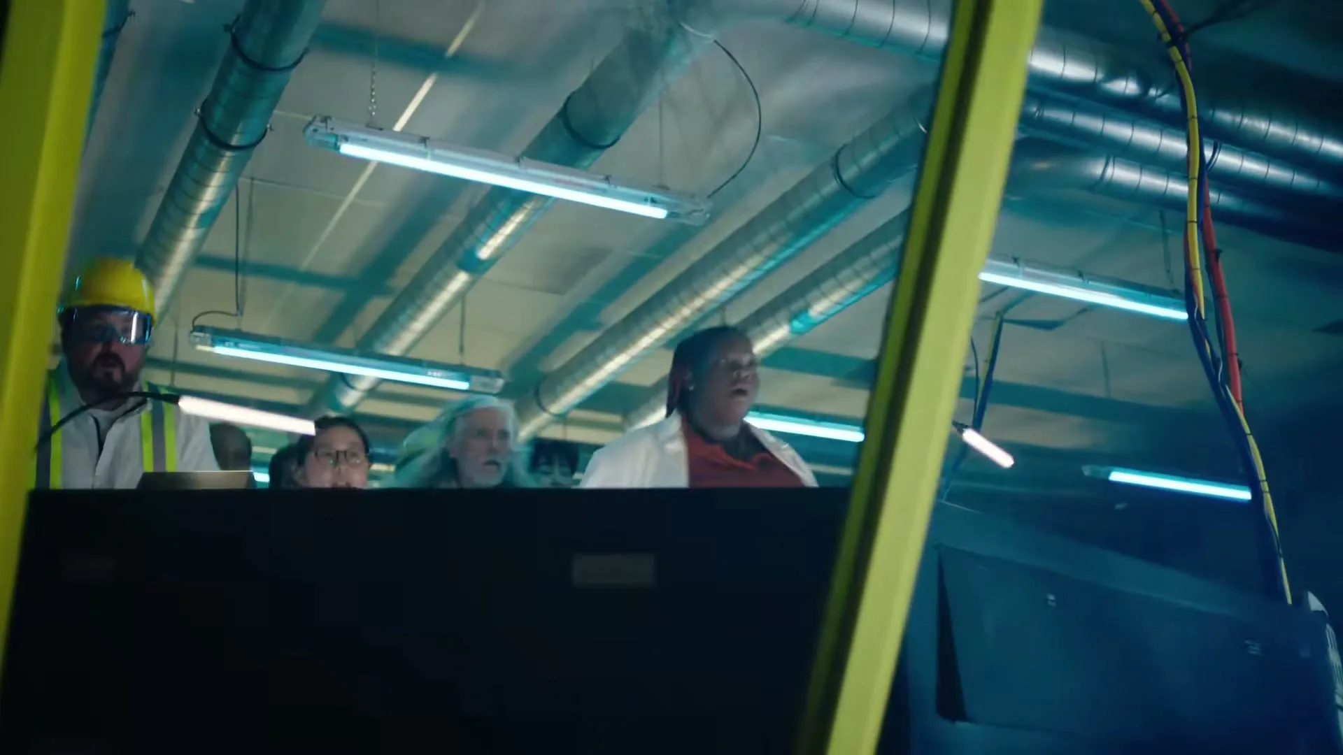 A diverse group of scientists and engineers look up in shock and awe in a futuristic, industrial control room with neon lights.