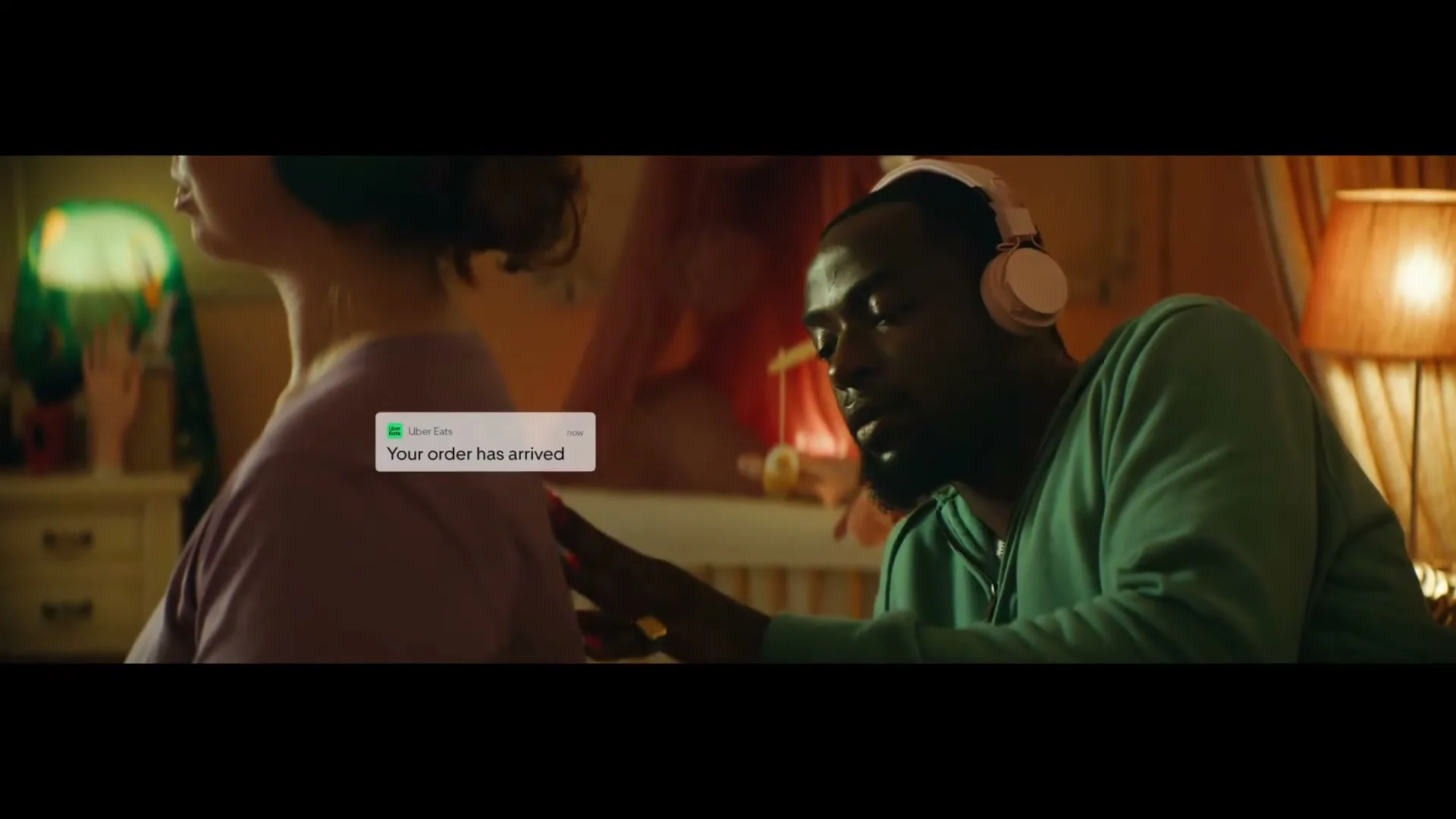 A young woman with green headphones sits with her back to the camera, while a young man with pink headphones touches her shoulder in a cozy room.