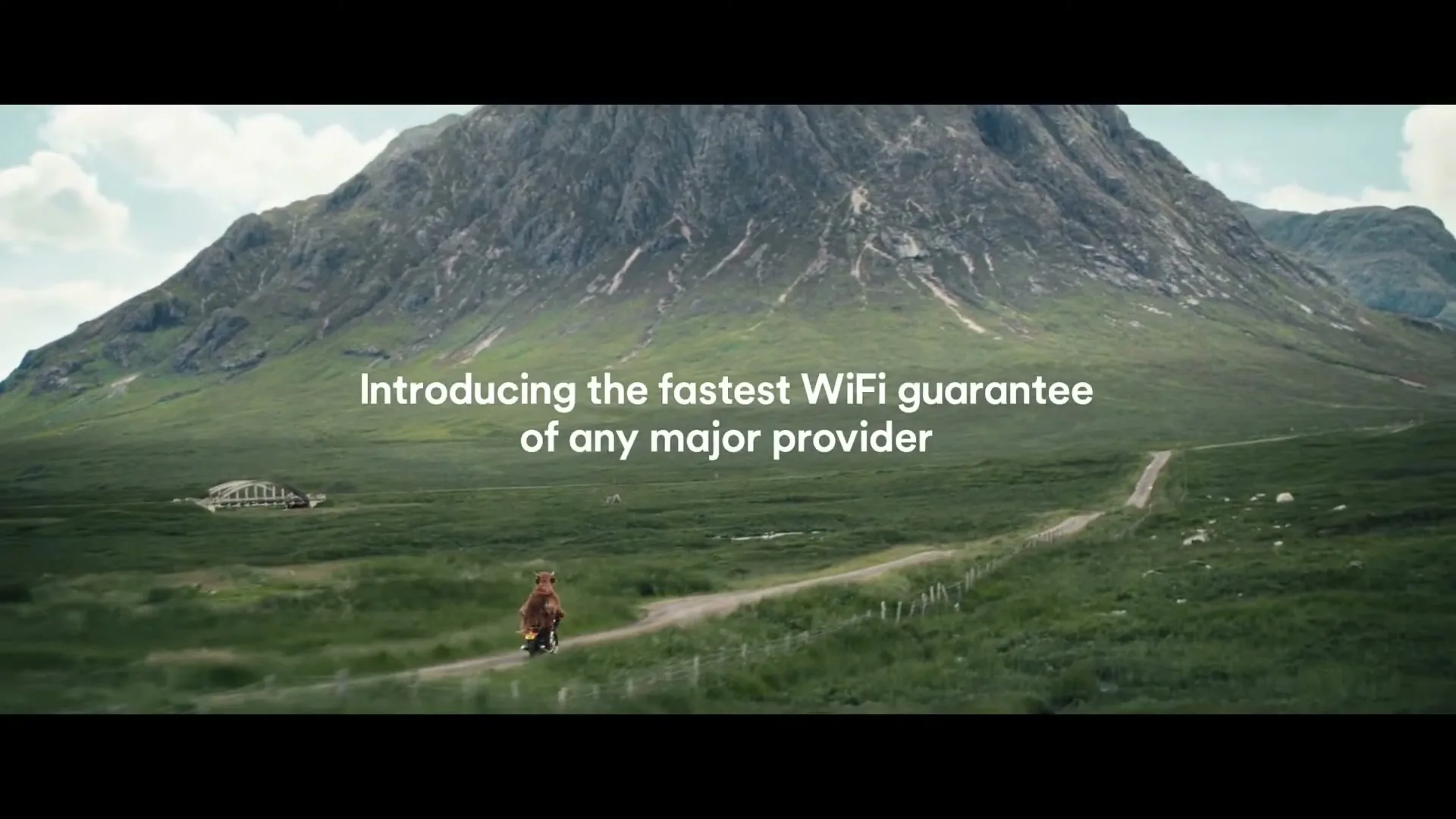 A shaggy brown Highland cow rides a red motorcycle down a dirt road in a vast green valley with mountains in the background, as text appears on screen.