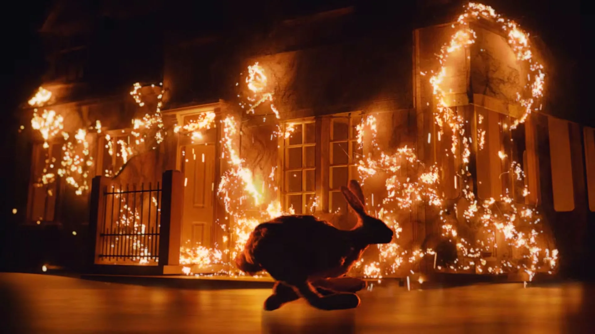 A rabbit-like creature, made of a flammable material, runs away from a burning house, engulfed in flames, at night.