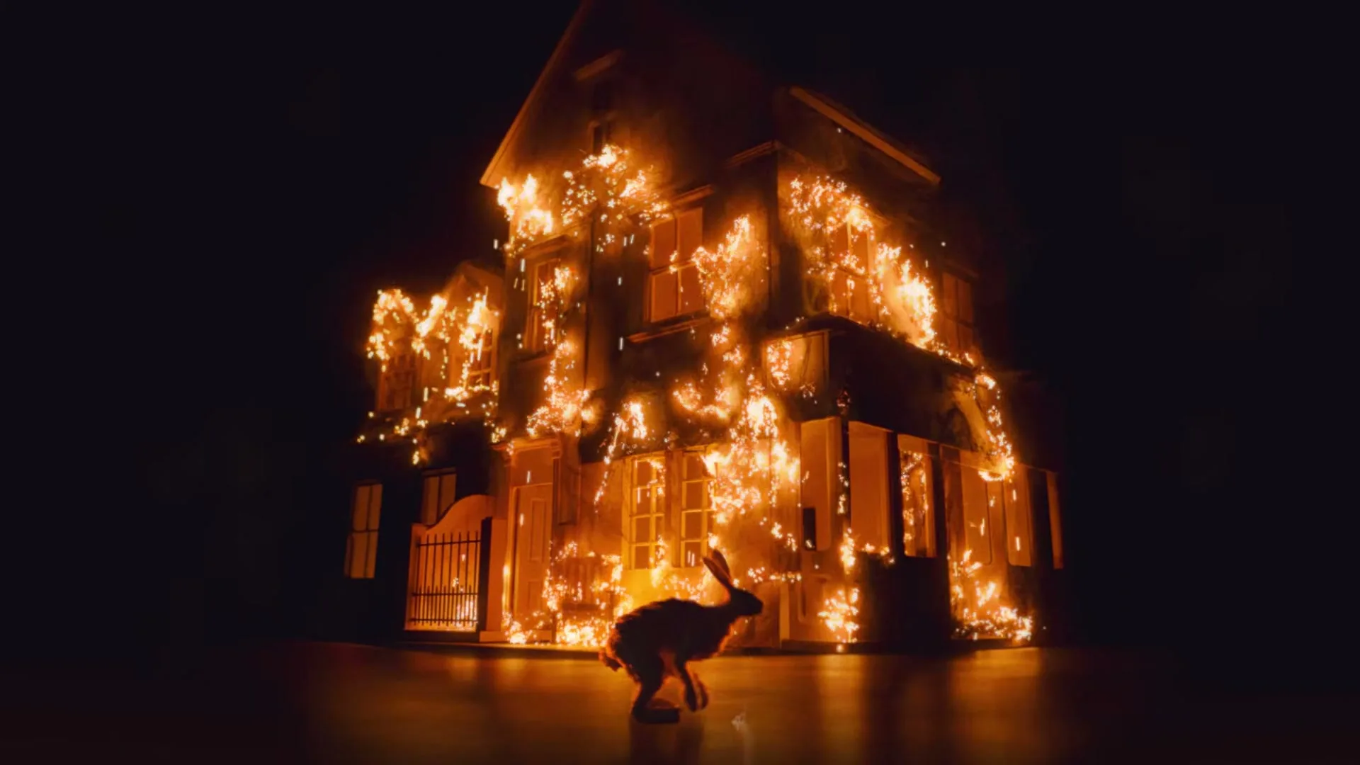 A rabbit runs past a burning miniature house, engulfed in flames, against a dark background, symbolizing destruction and loss.