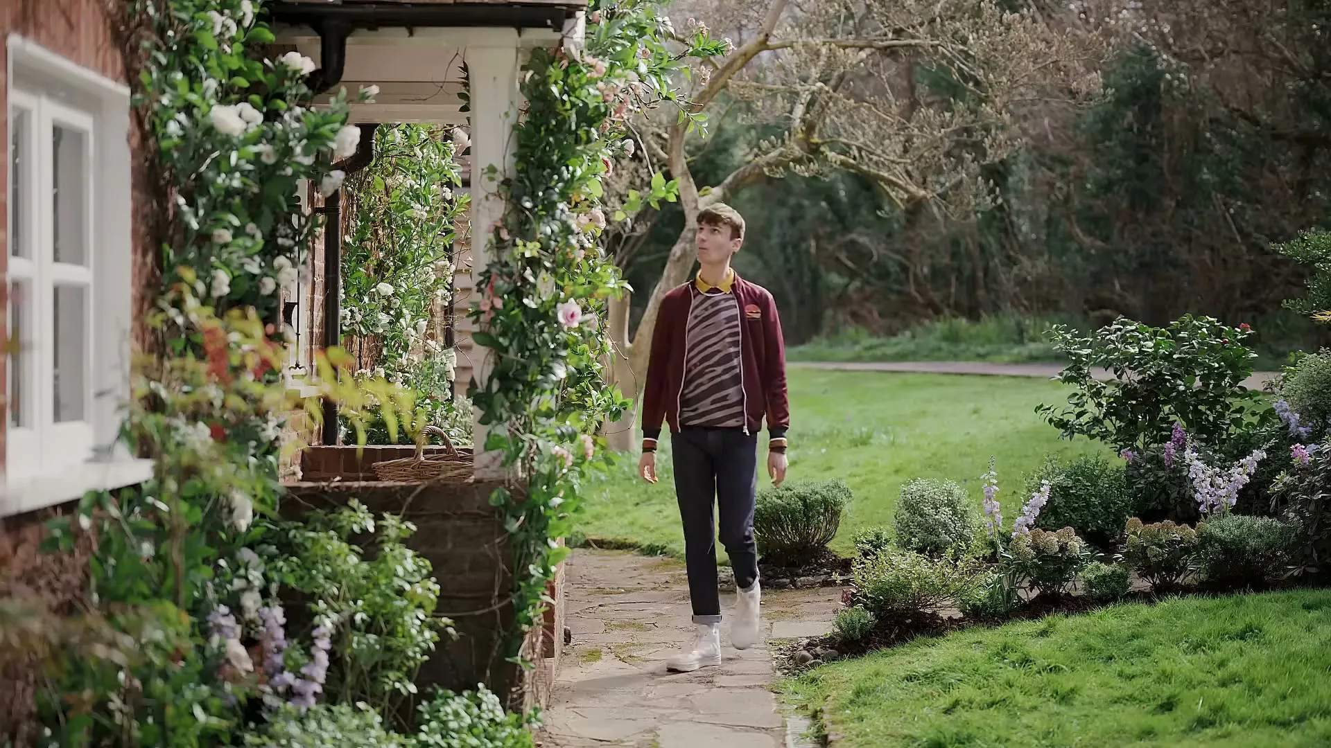 A young man in a striped shirt and maroon jacket walks along a stone path next to a house covered in climbing plants, looking around.