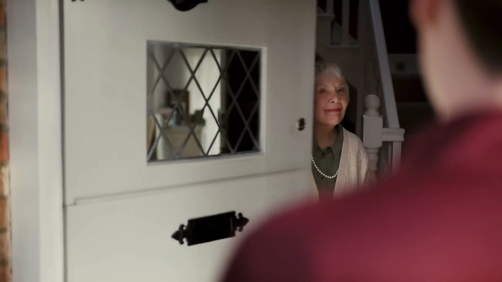 A man in a red shirt stands at a white front door, which opens to reveal an elderly woman smiling inside her home.