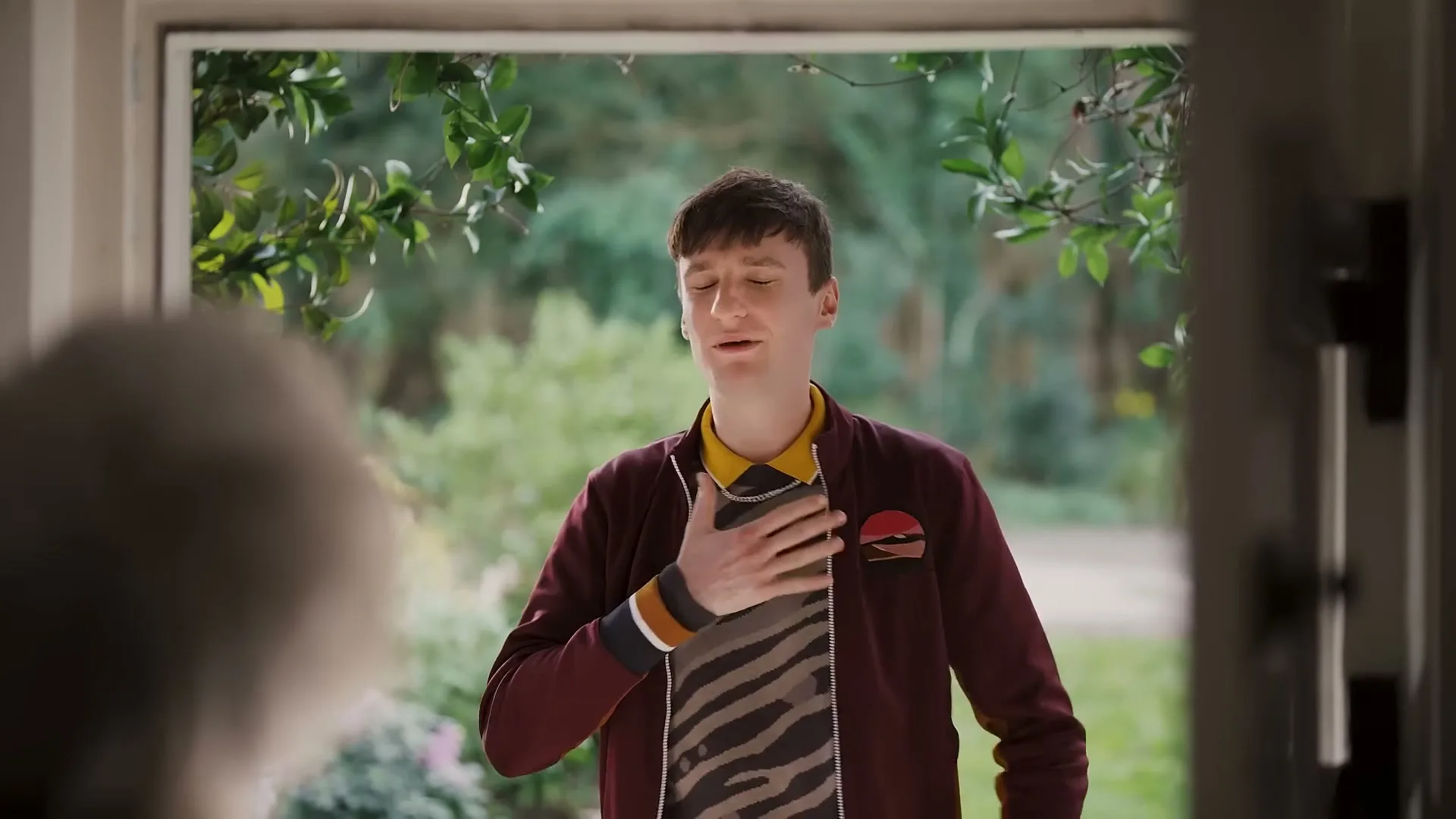 A young man in a bomber jacket and patterned shirt stands at a doorway, talking to someone off-screen, with a green garden visible behind him.