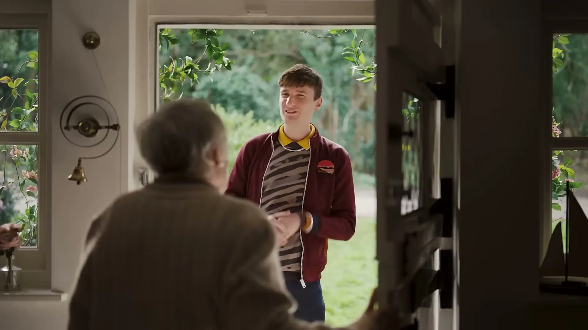 A young man with a striped shirt and a maroon jacket stands at an open doorway, smiling at an older woman whose back is to the camera, in a suburban home.