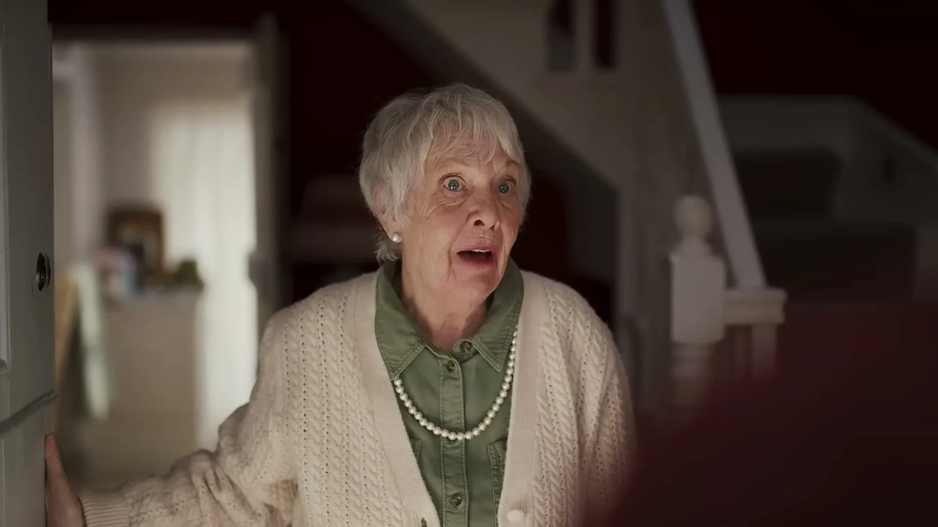 An elderly woman with white hair and a pearl necklace stands in a doorway, looking surprised and then smiling, in a dimly lit house interior.