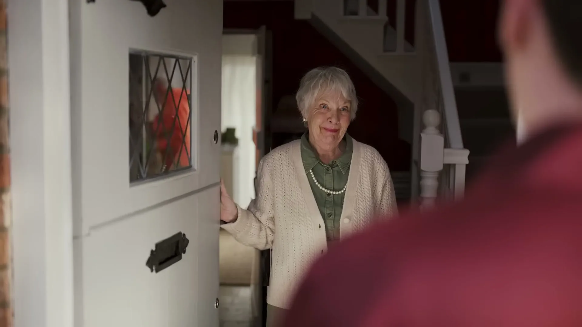 An elderly woman with white hair and a pearl necklace opens her front door, looking surprised at the person standing outside.