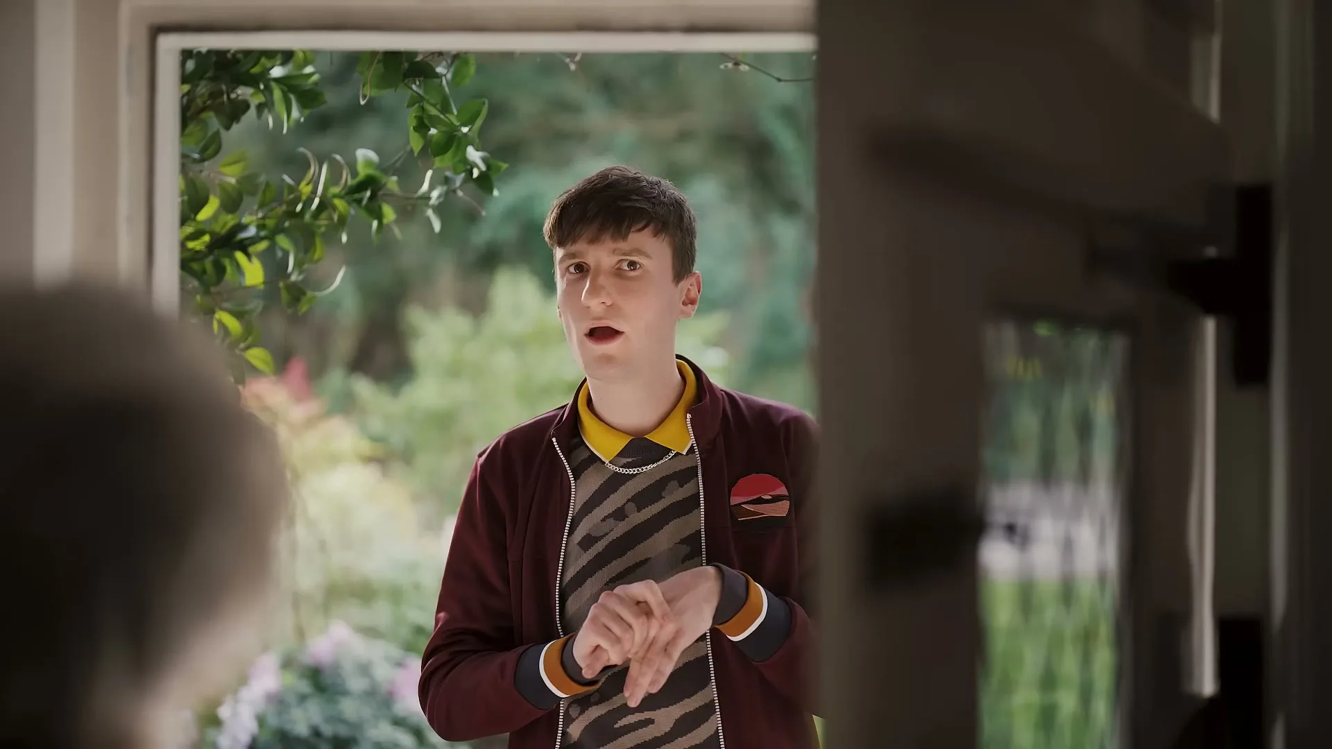 A young man in a maroon jacket and patterned sweater stands at a doorway, looking surprised, as seen from inside a house.