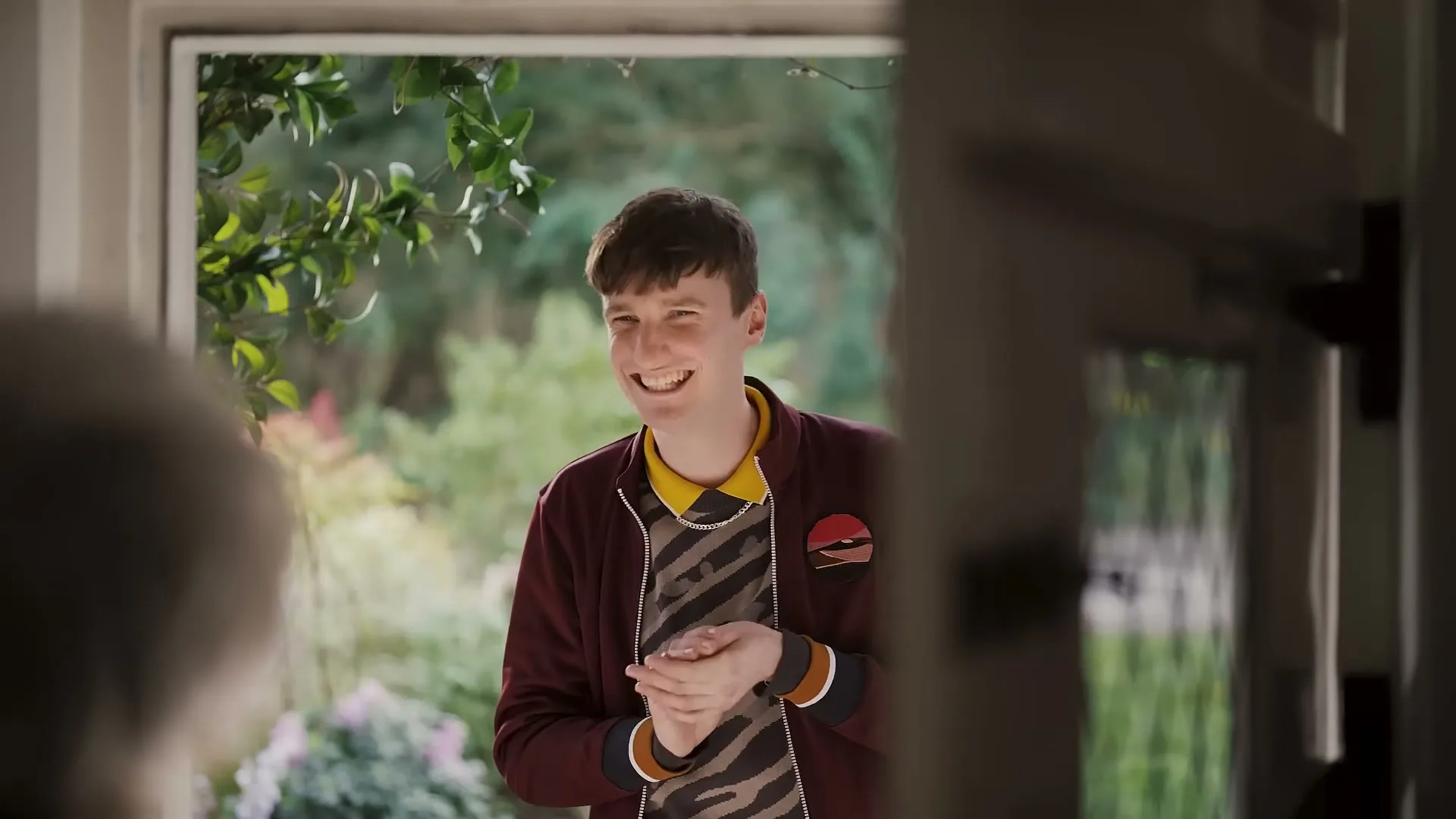A young man with short brown hair, wearing a maroon jacket and a patterned shirt, stands smiling at a doorway.
