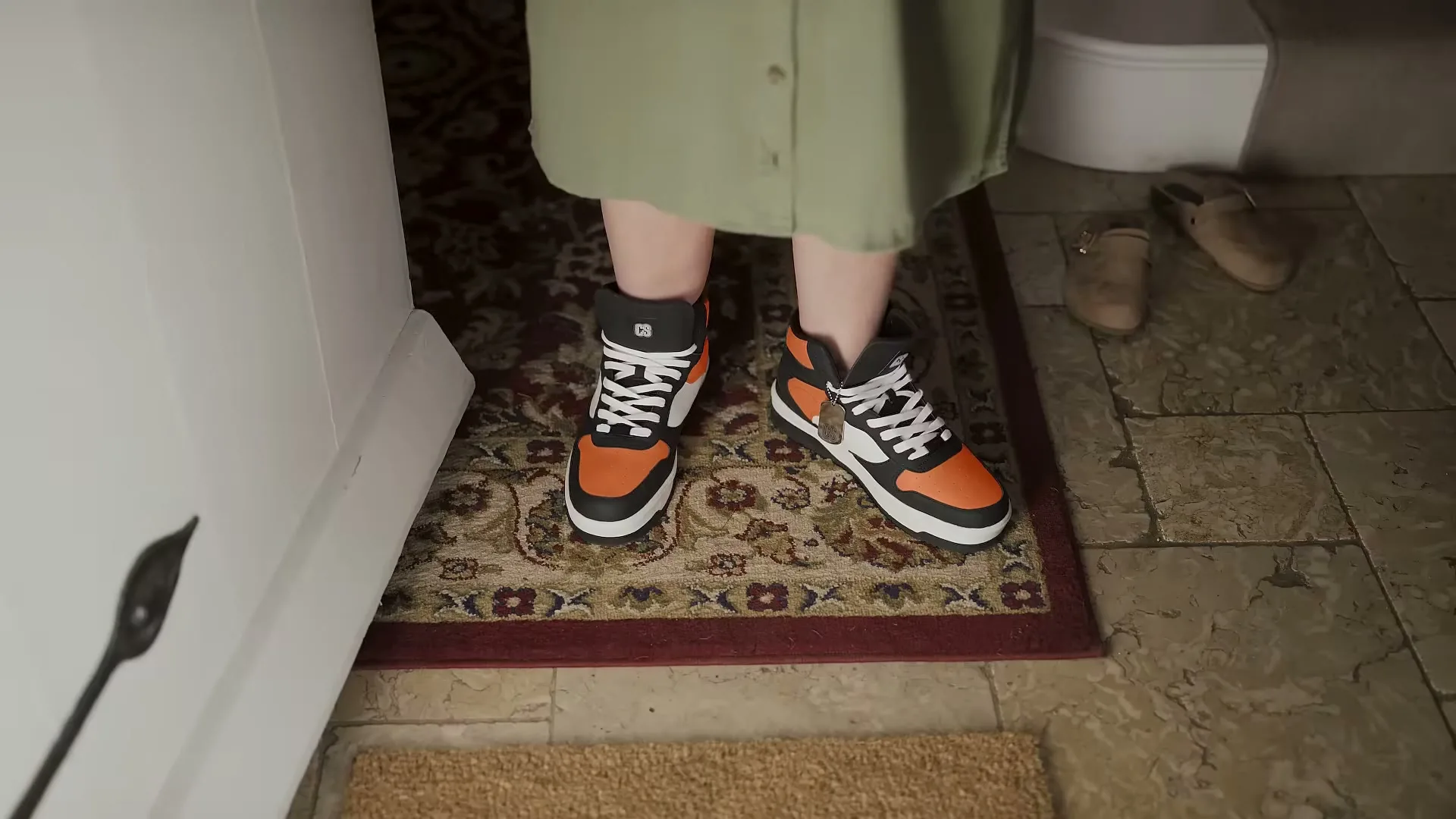 A person wearing a green dress and orange and black sneakers stands on a patterned rug inside a house, with other shoes visible nearby.