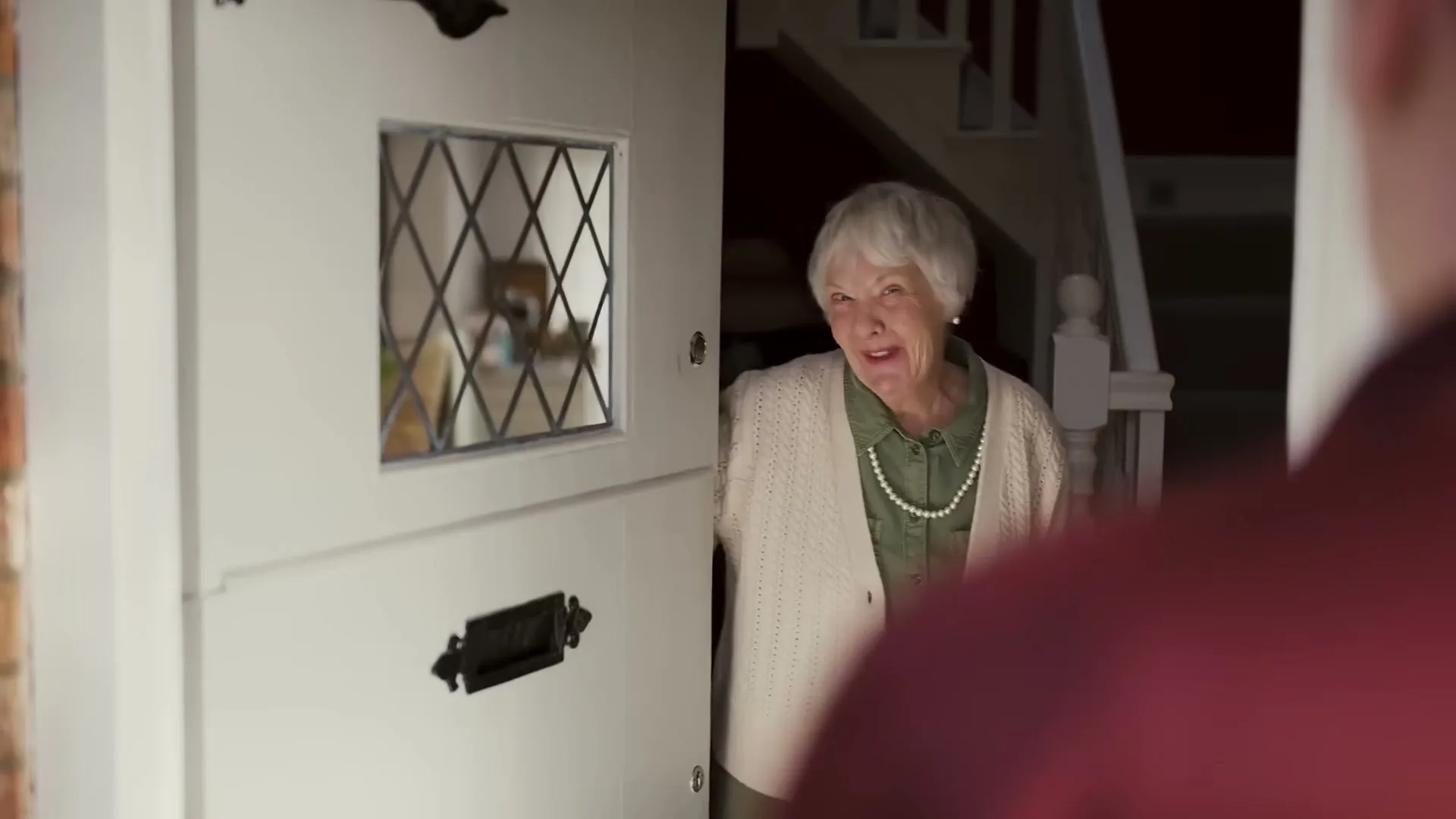 An elderly woman with white hair and a pearl necklace opens her front door, smiling at someone off-camera.