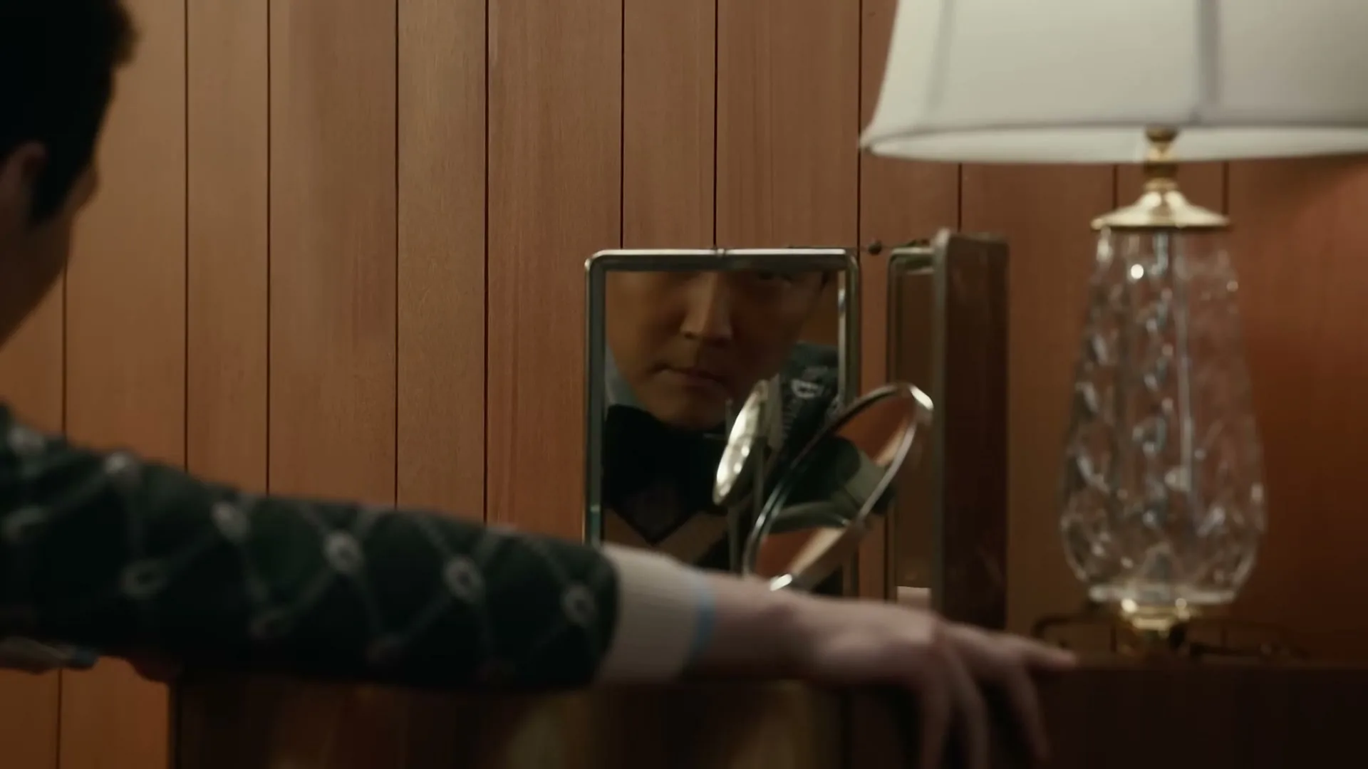 A man in a green sweater looks at his reflection in a small mirror on a wooden dresser in a warmly lit room.