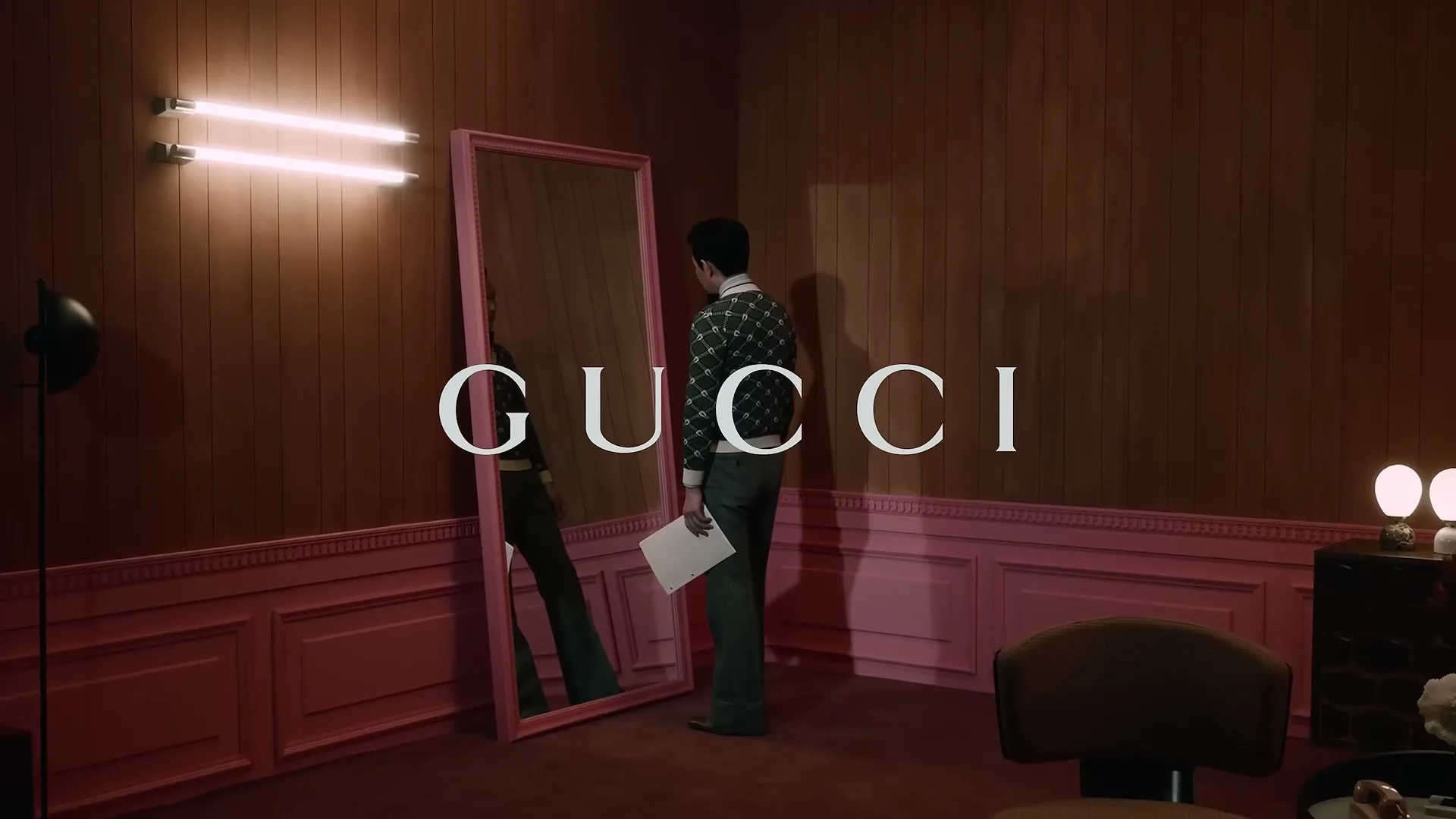 A man in a patterned sweater and green pants stands in front of a pink-framed mirror in a wood-paneled room with pink wainscoting, holding papers.