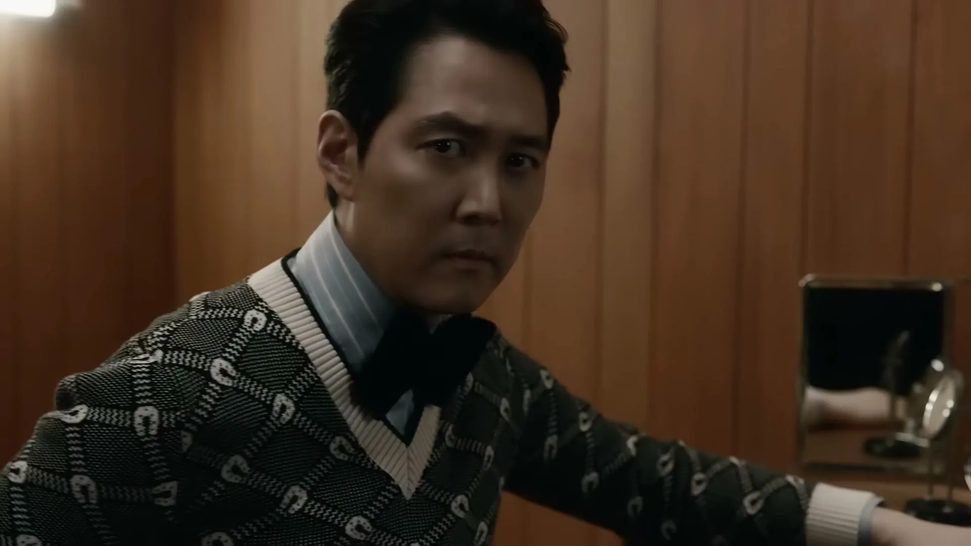 A man in a patterned sweater and bow tie looks intently at the camera in a wood-paneled room.