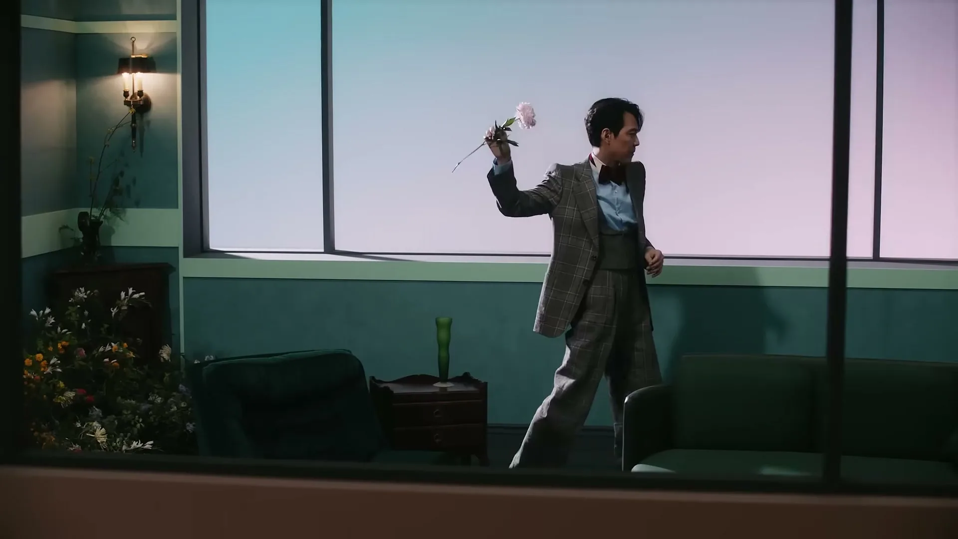 A man in a plaid suit and bow tie holds a pink flower, walking across a teal room with large windows.