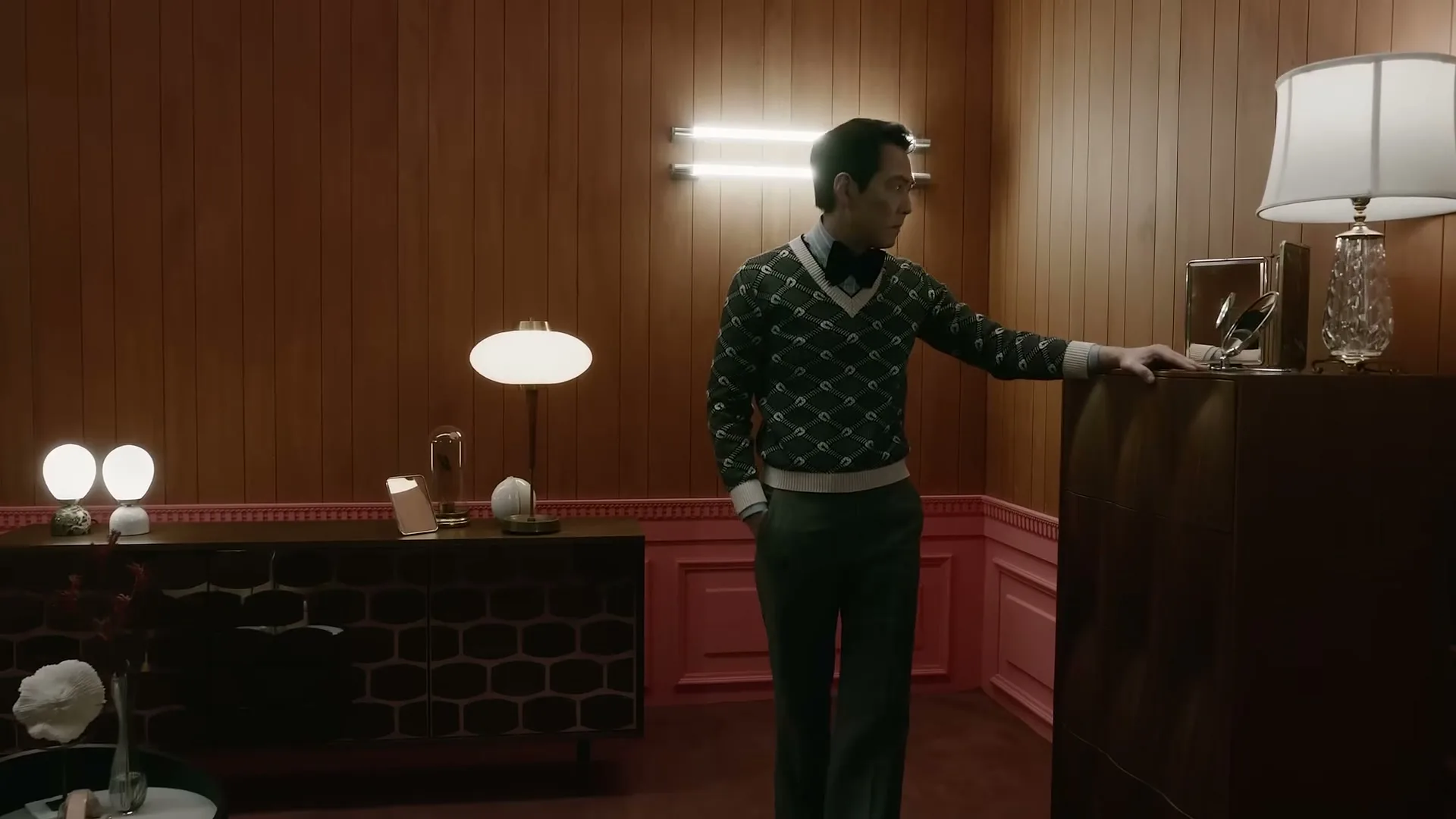 A man in a patterned sweater and bow tie stands in a wood-paneled room with pink trim, looking to the right.
