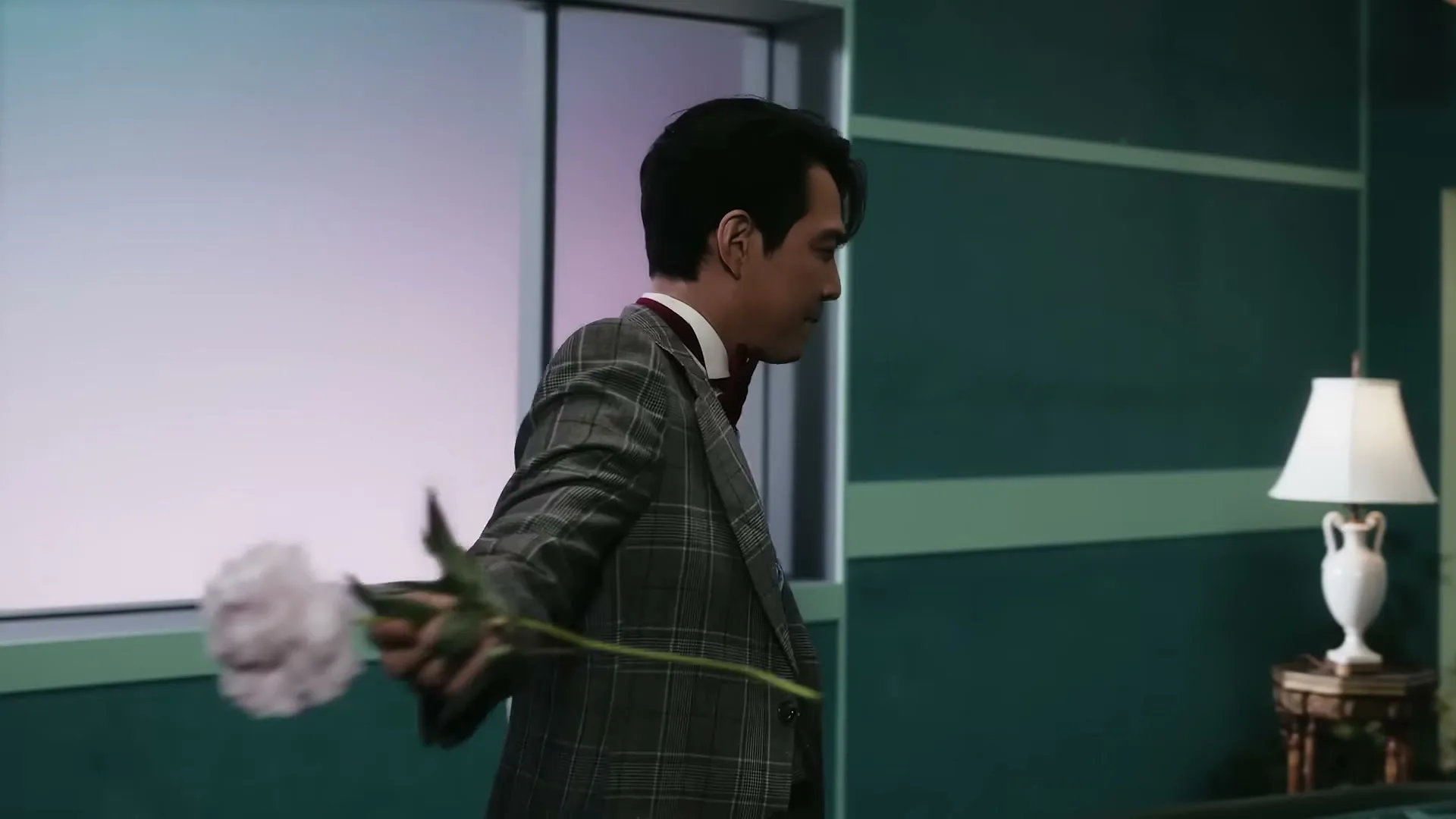 A man in a plaid suit and bow tie walks through a room, holding a pink flower in his outstretched hand.