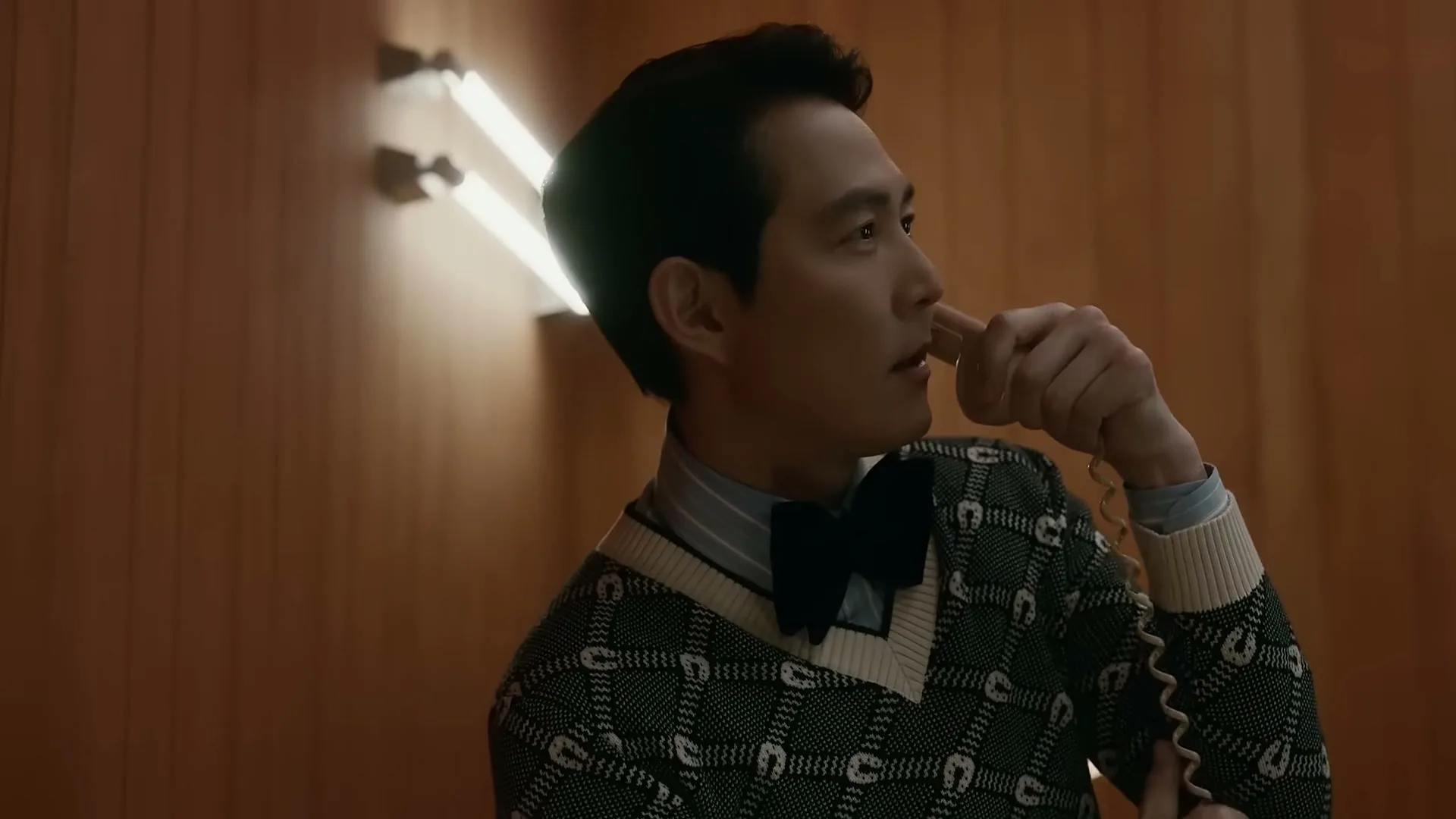 A man in a patterned sweater and bow tie holds a phone receiver to his mouth, looking off-camera in a wood-paneled room.