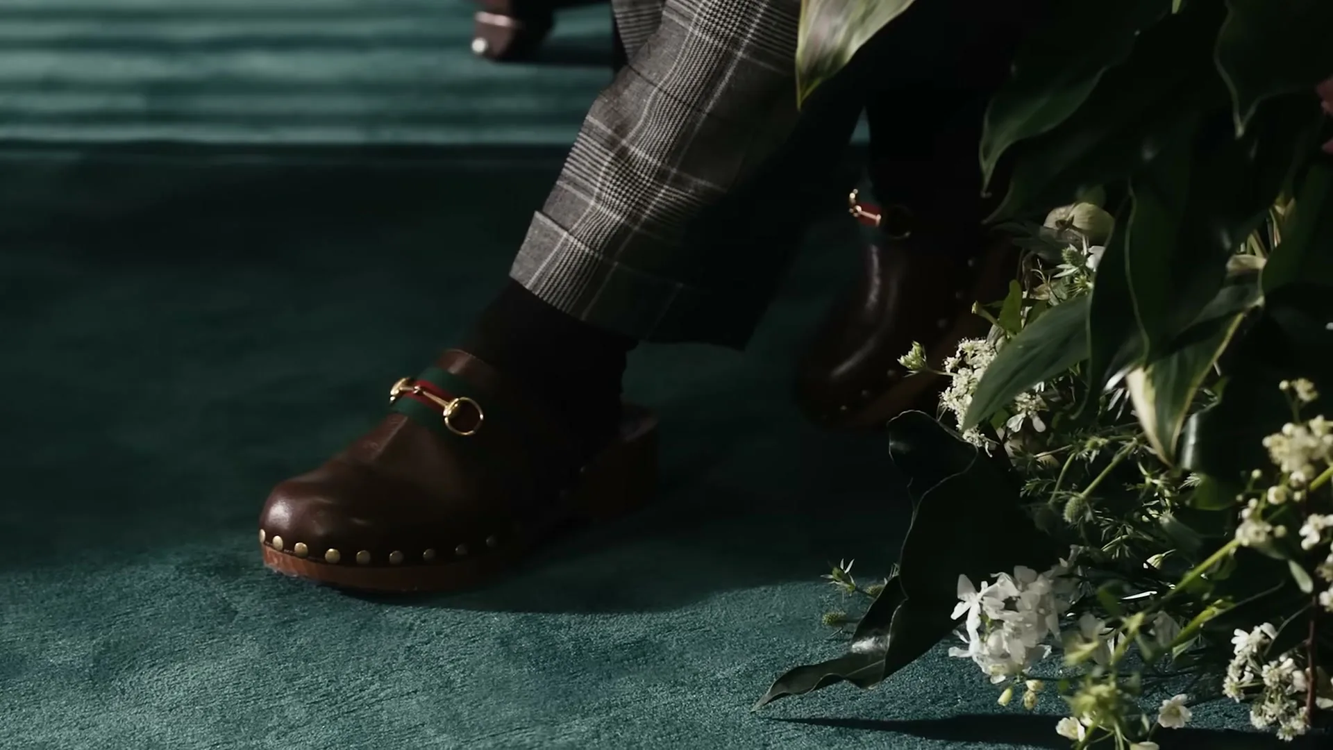 A person wearing plaid pants and brown clogs with a gold buckle and green and red stripe sits on a teal carpet next to a bouquet of white flowers.
