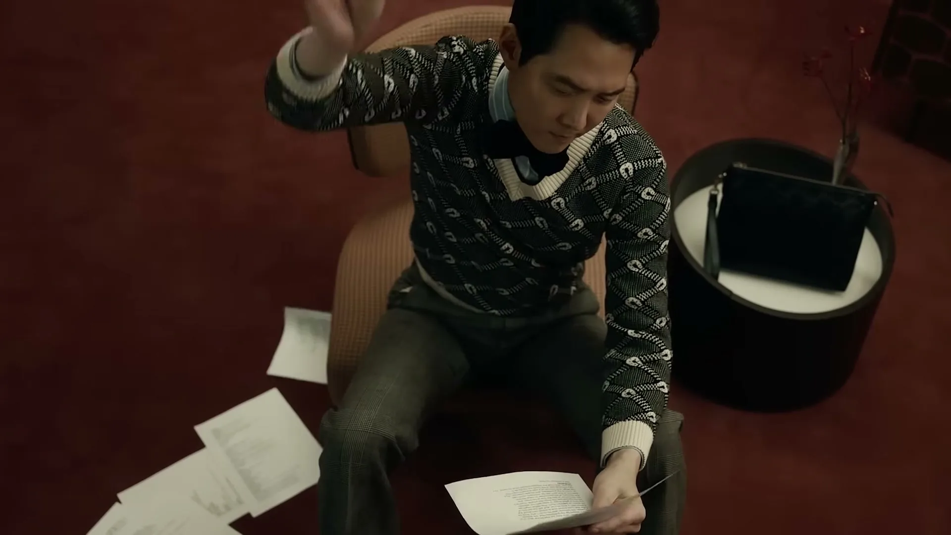 A man in a patterned sweater sits on a chair, looking frustrated as he throws papers around a luxurious room.