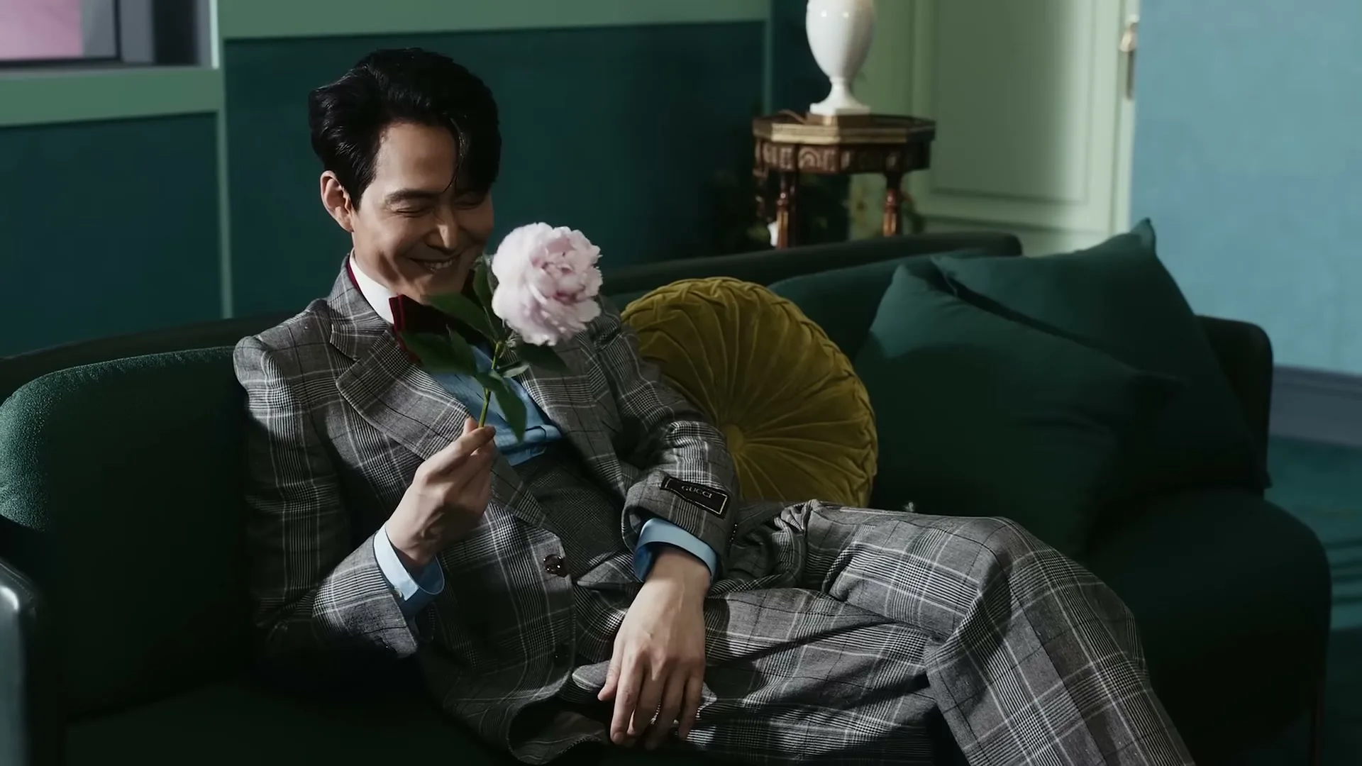 A man in a plaid suit reclines on a green couch, smiling as he holds a pink flower to his nose in a luxurious room.