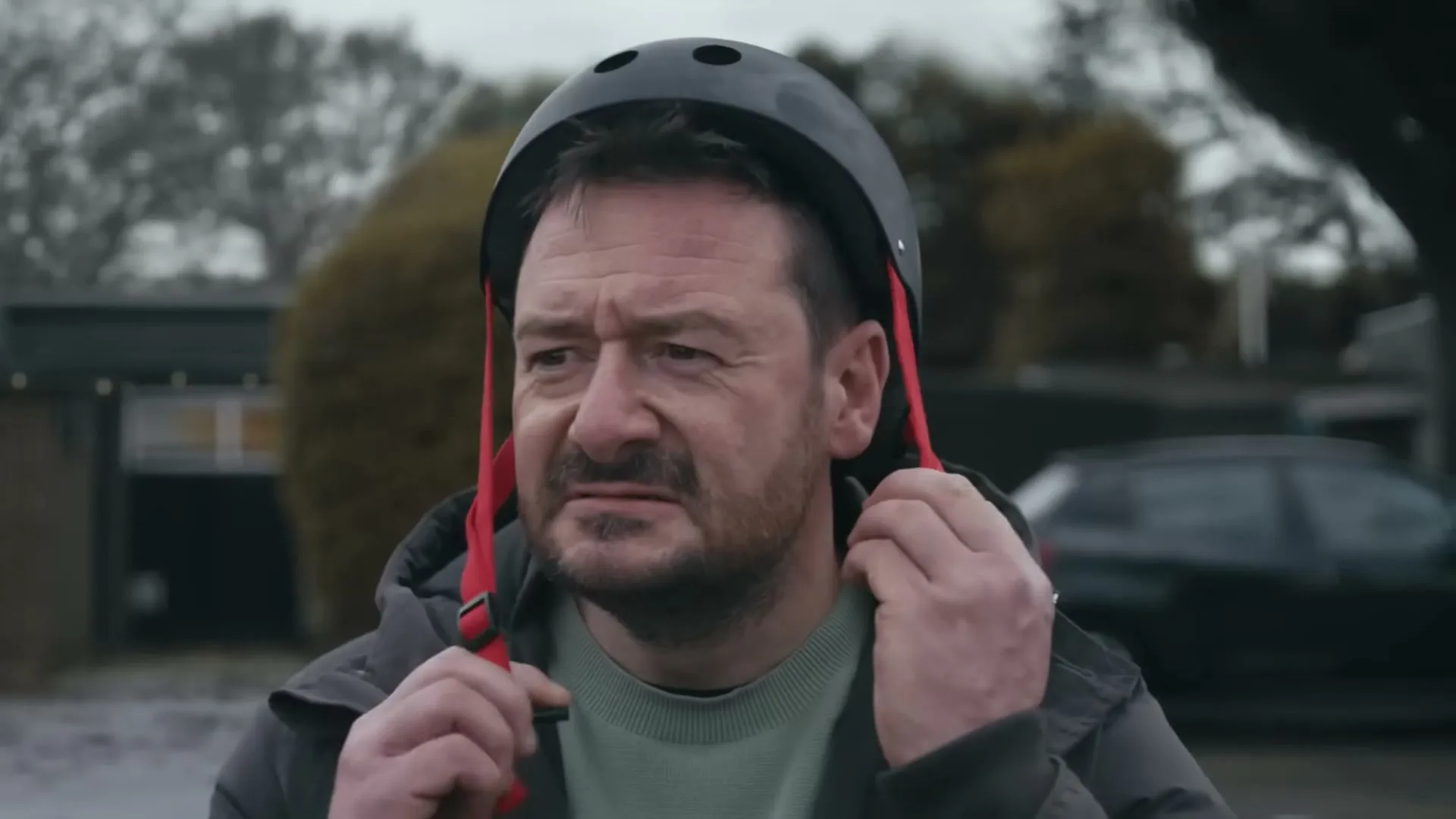 A man in his 40s with a beard and a dark jacket puts on a black helmet with red straps, looking determined in a suburban street.