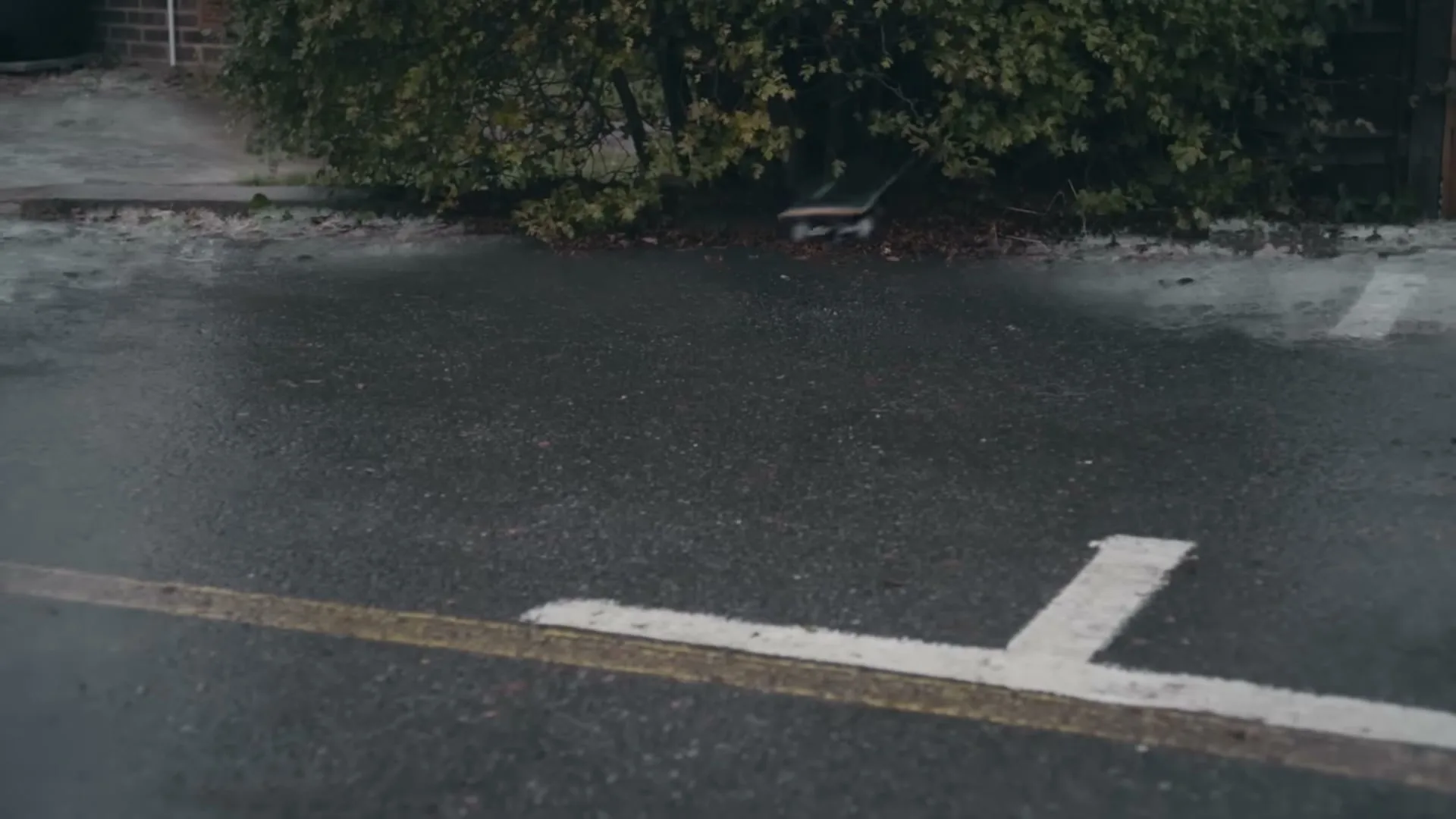 A skateboard rolls across a wet asphalt road and crashes into a bush on the side of the road.