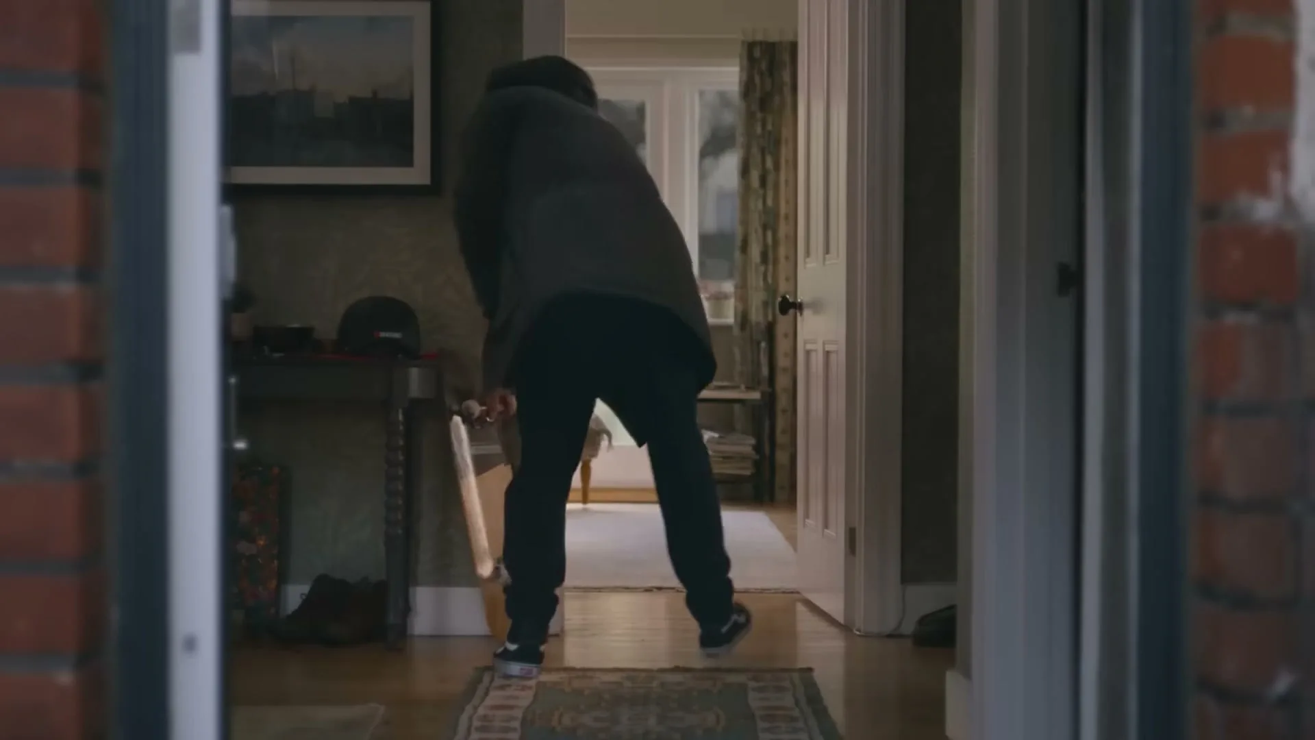 A man in a dark coat and jeans walks into a house, holding a skateboard and a helmet, in a medium shot.