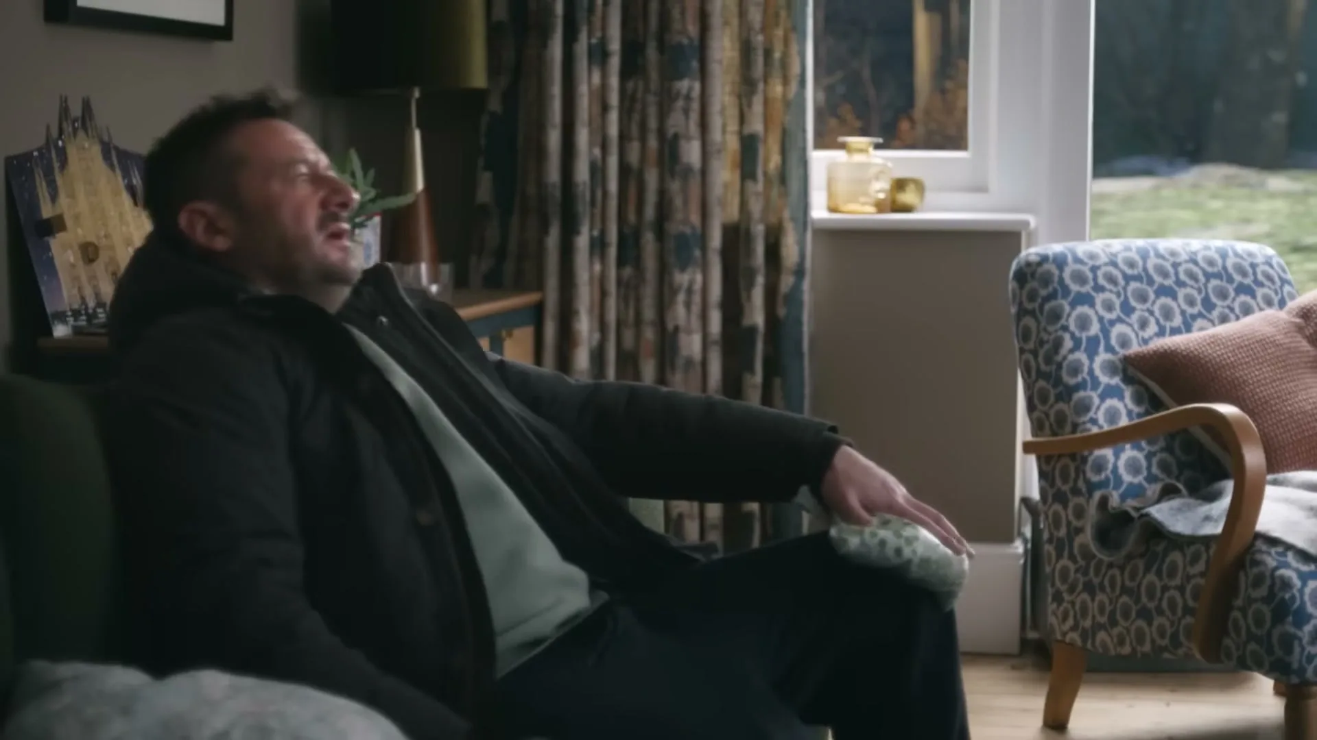 A man in his 40s, wearing a dark jacket and light green t-shirt, sits on a green sofa, holding an ice pack to his knee, wincing in pain in a living room.