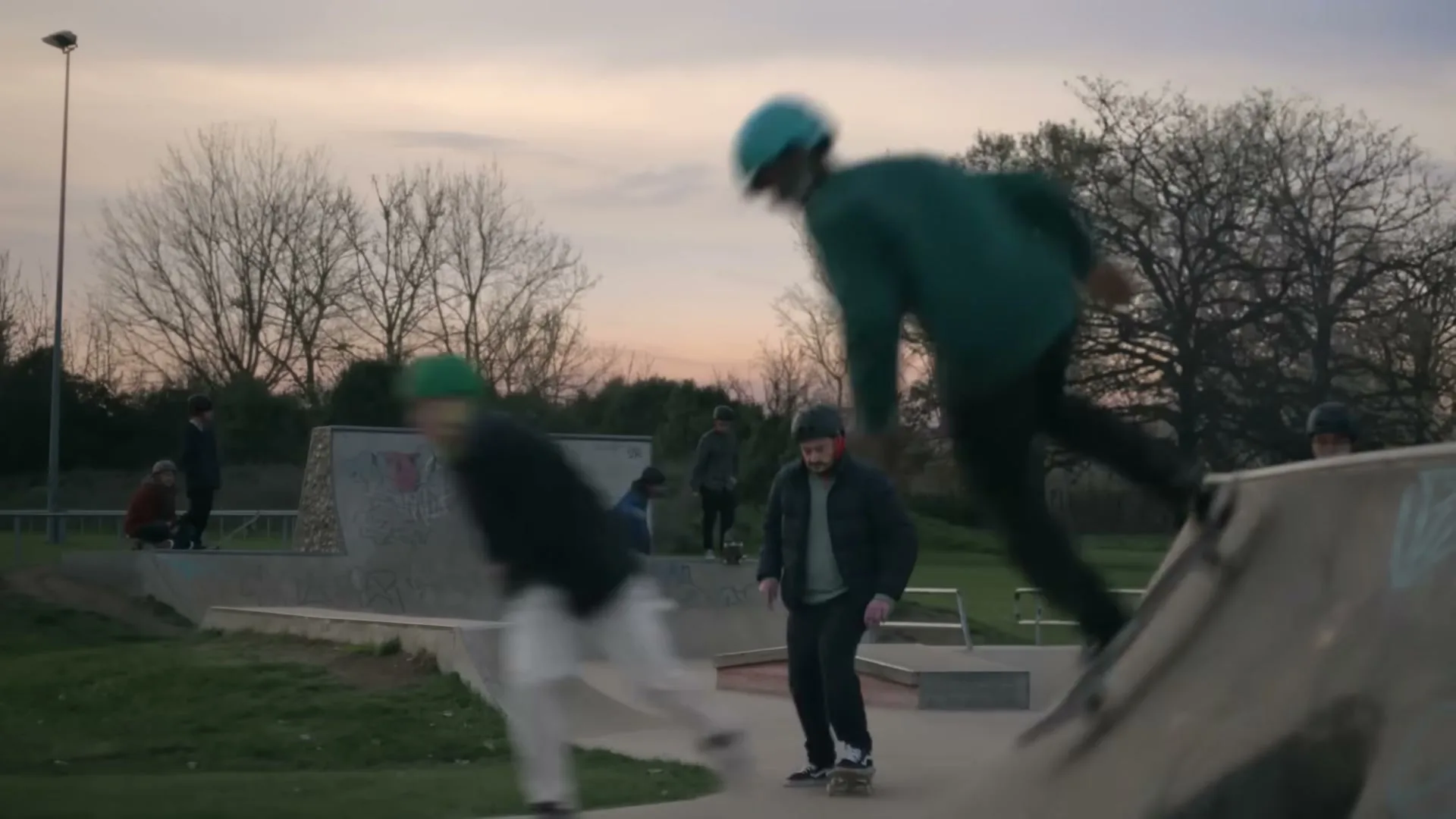 A man in his 30s attempts to skateboard in a concrete skatepark, while other people skate and watch in the background at dusk.