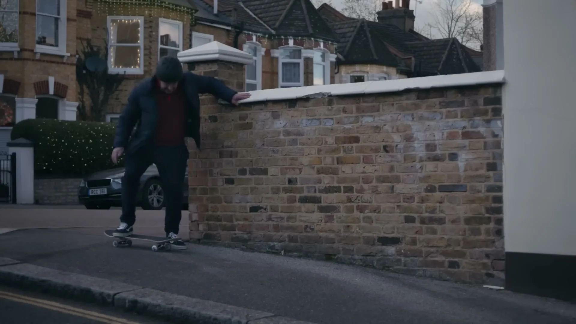 A man in a dark jacket and red sweater attempts to skateboard on a suburban street, holding onto a brick wall for balance.