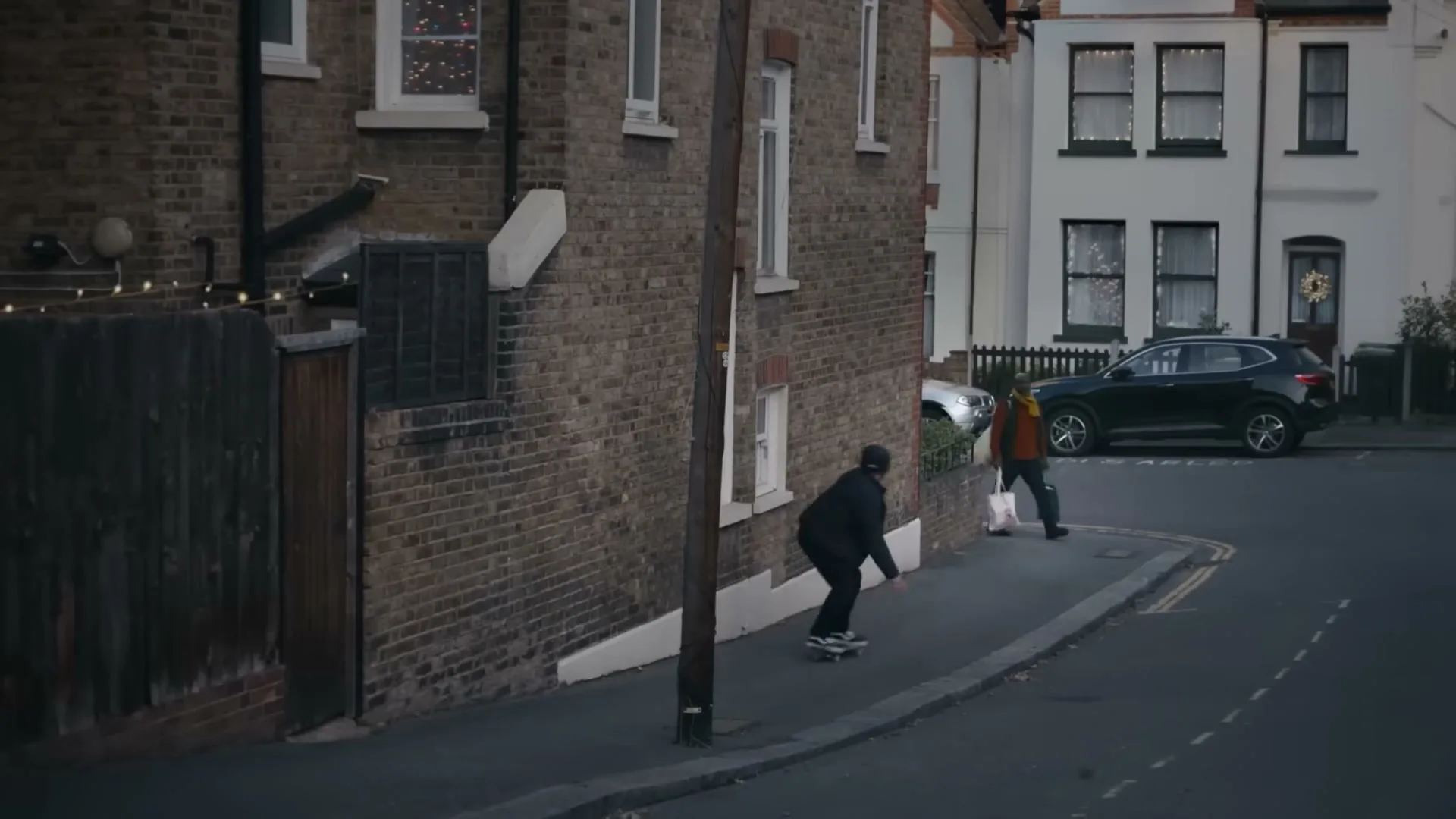 A young man in a black hoodie and cap attempts to skateboard down a suburban street, losing his balance and falling as another person walks by.