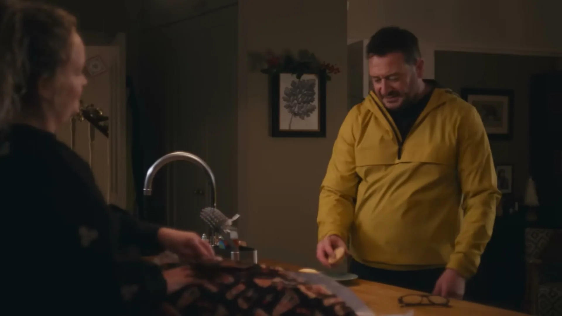A man in a yellow jacket stands in a kitchen, looking at a woman who is wrapping a gift on the counter.