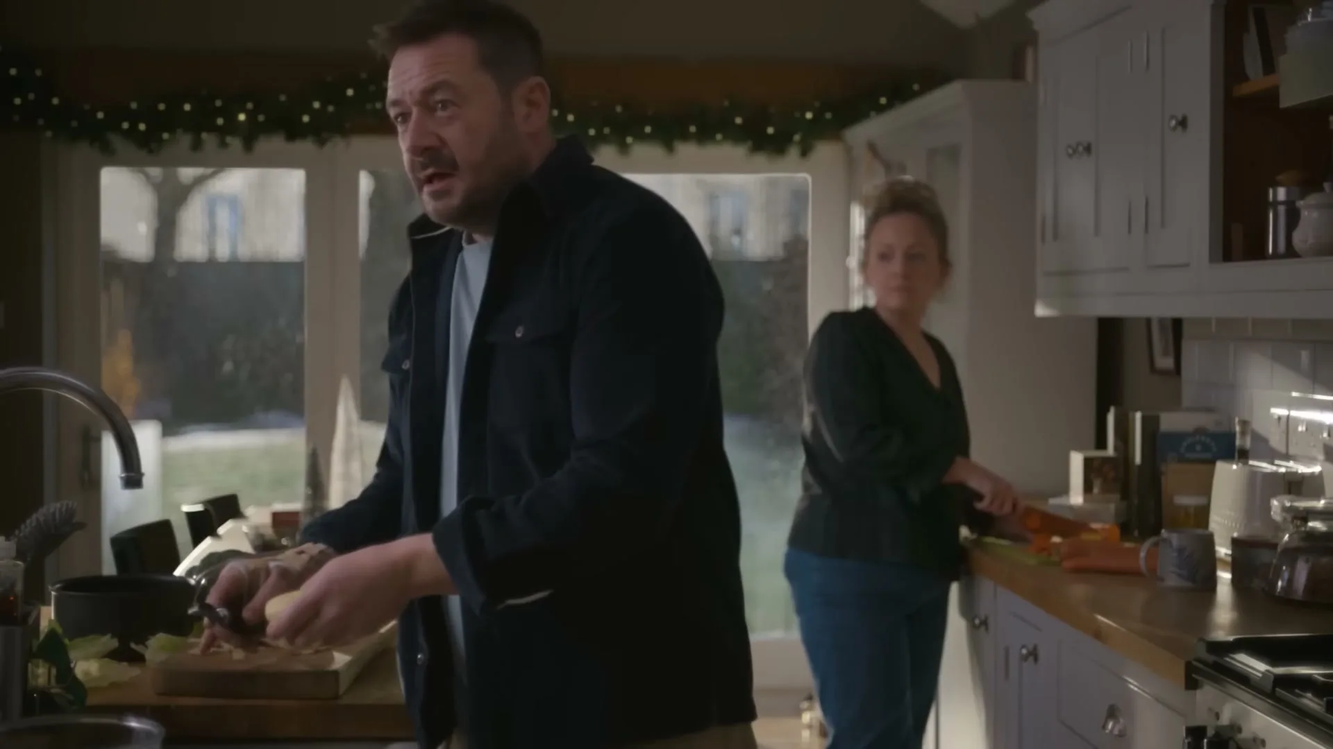 A man and a woman are in a kitchen, preparing food. The man is peeling potatoes at a sink, and the woman is chopping vegetables on a counter.
