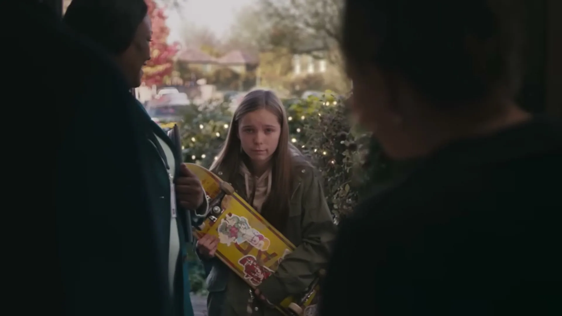 A young girl with long brown hair, wearing a green jacket, stands awkwardly holding a skateboard with stickers, while an adult woman smiles beside her at a doorway.