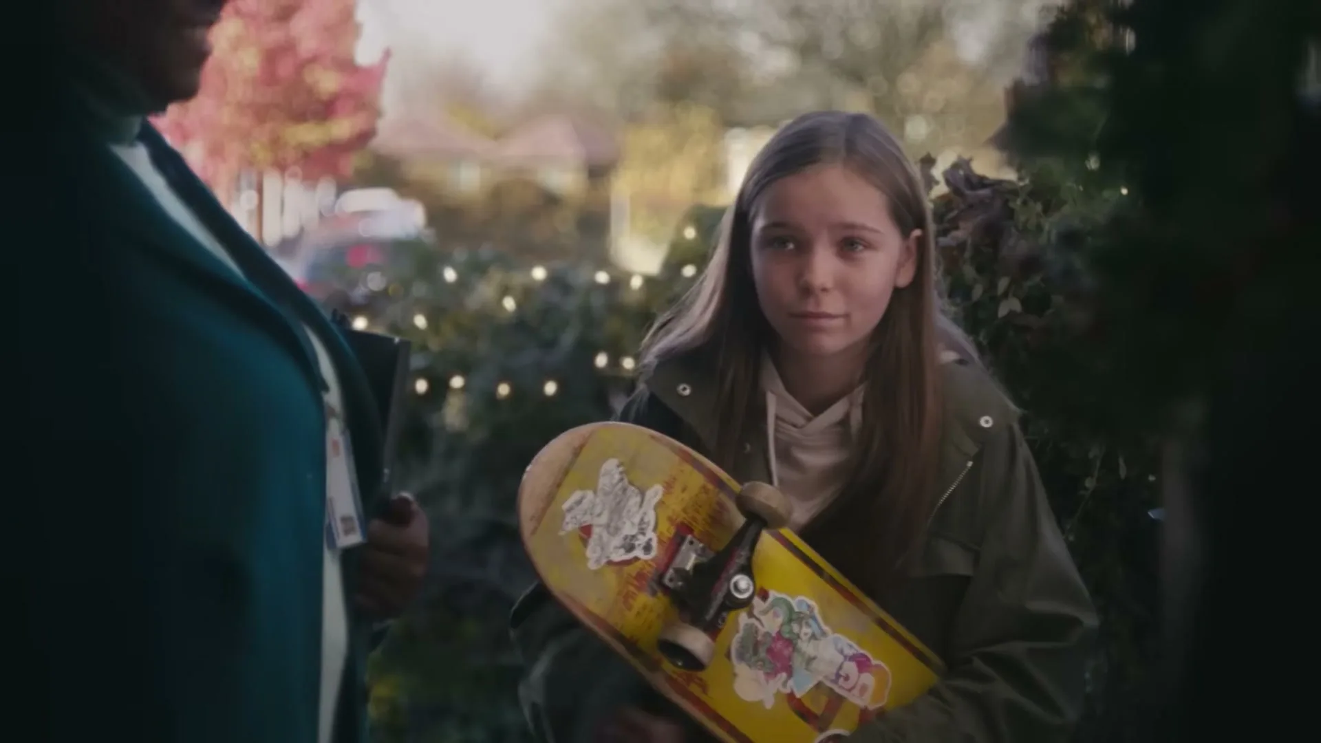 A young girl with long brown hair, wearing a green jacket, holds a yellow skateboard and looks at someone off-screen at a house entrance.