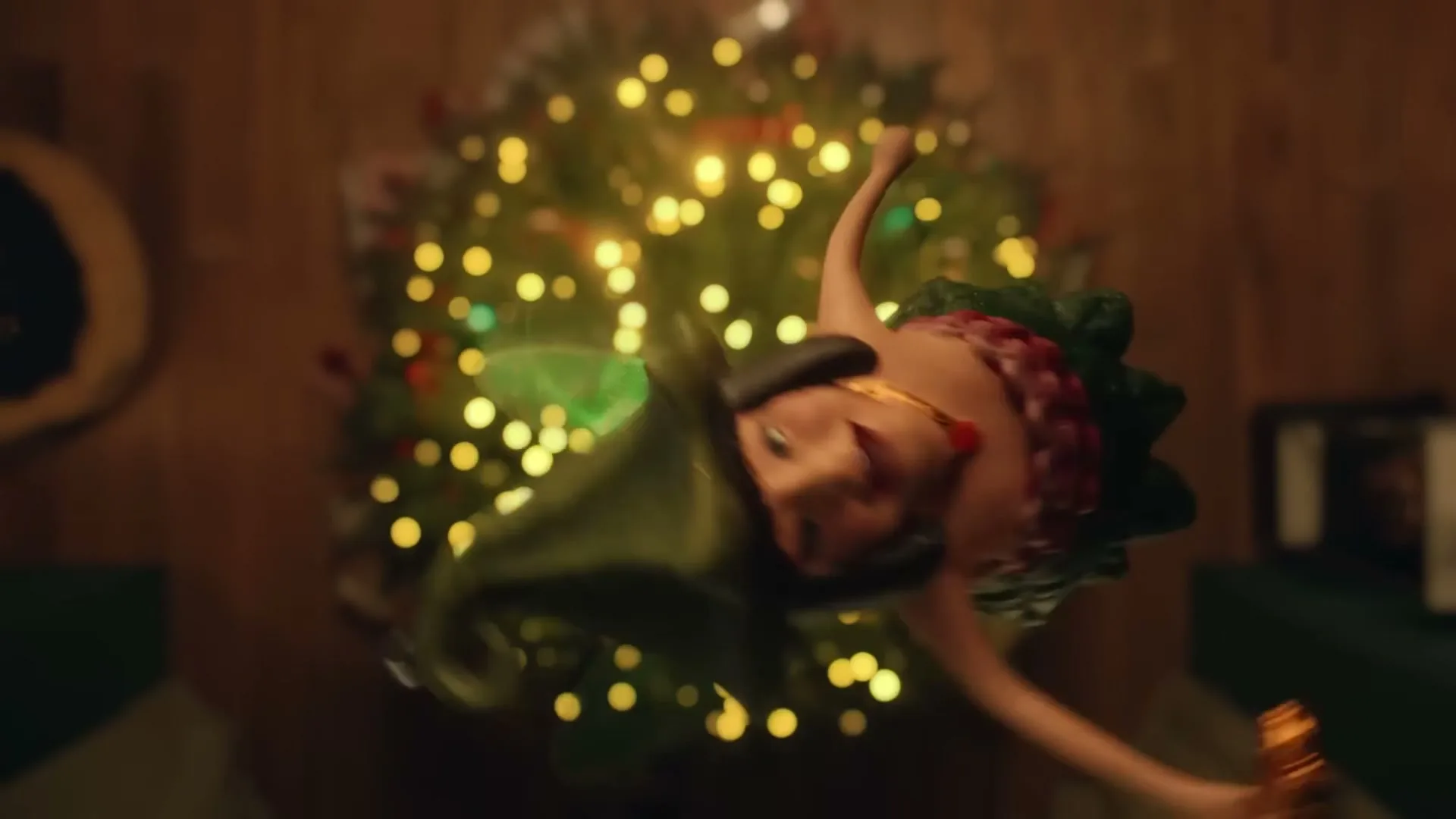 A CGI Christmas fairy, wearing a green and red dress, flies around a decorated Christmas tree in a cozy living room.