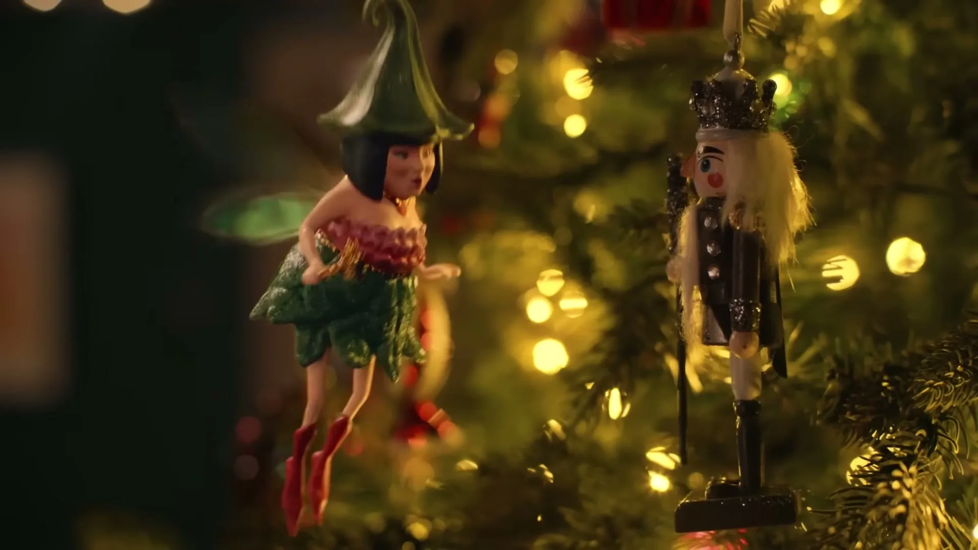 A small, green-clad fairy ornament with wings swings on a Christmas tree, looking at a nutcracker ornament, surrounded by warm lights.