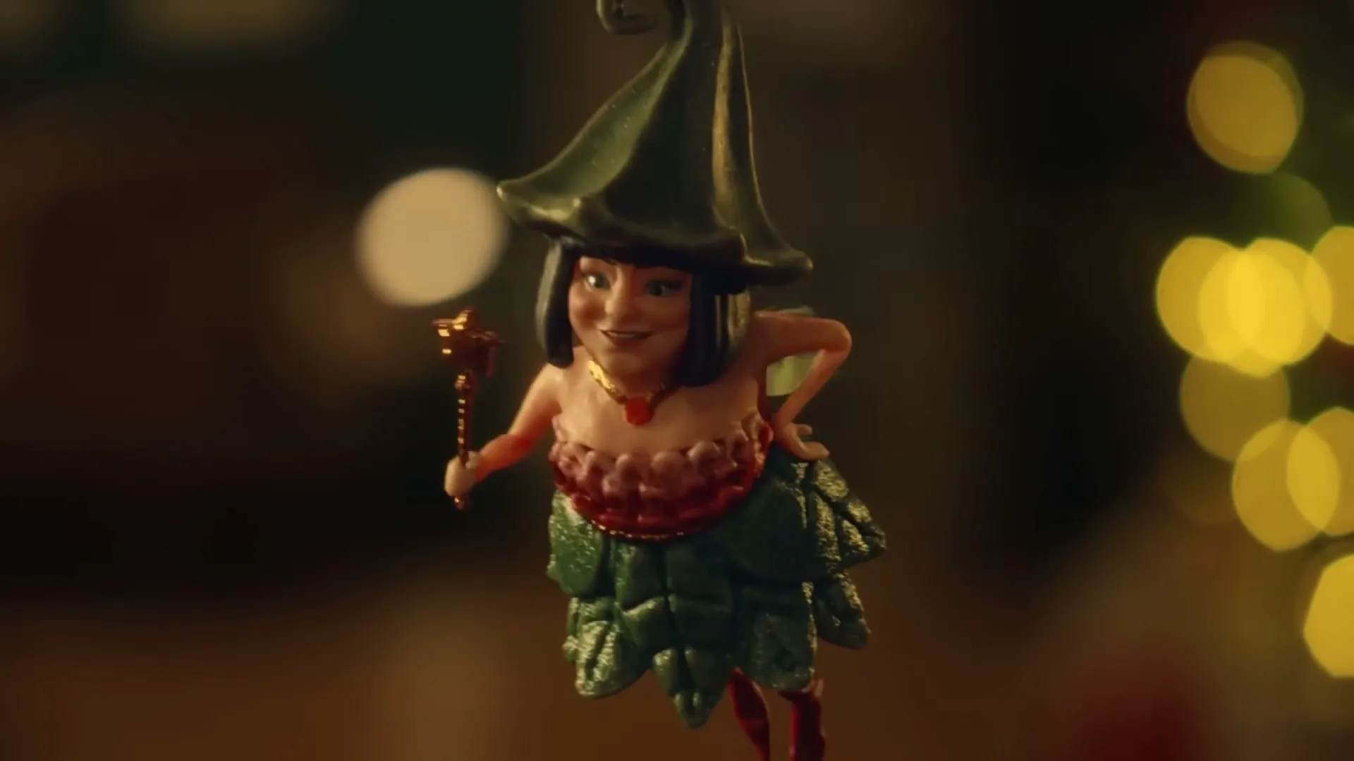 A small, green-hatted fairy ornament with a red top and green skirt hangs on a Christmas tree, holding a star-tipped wand.