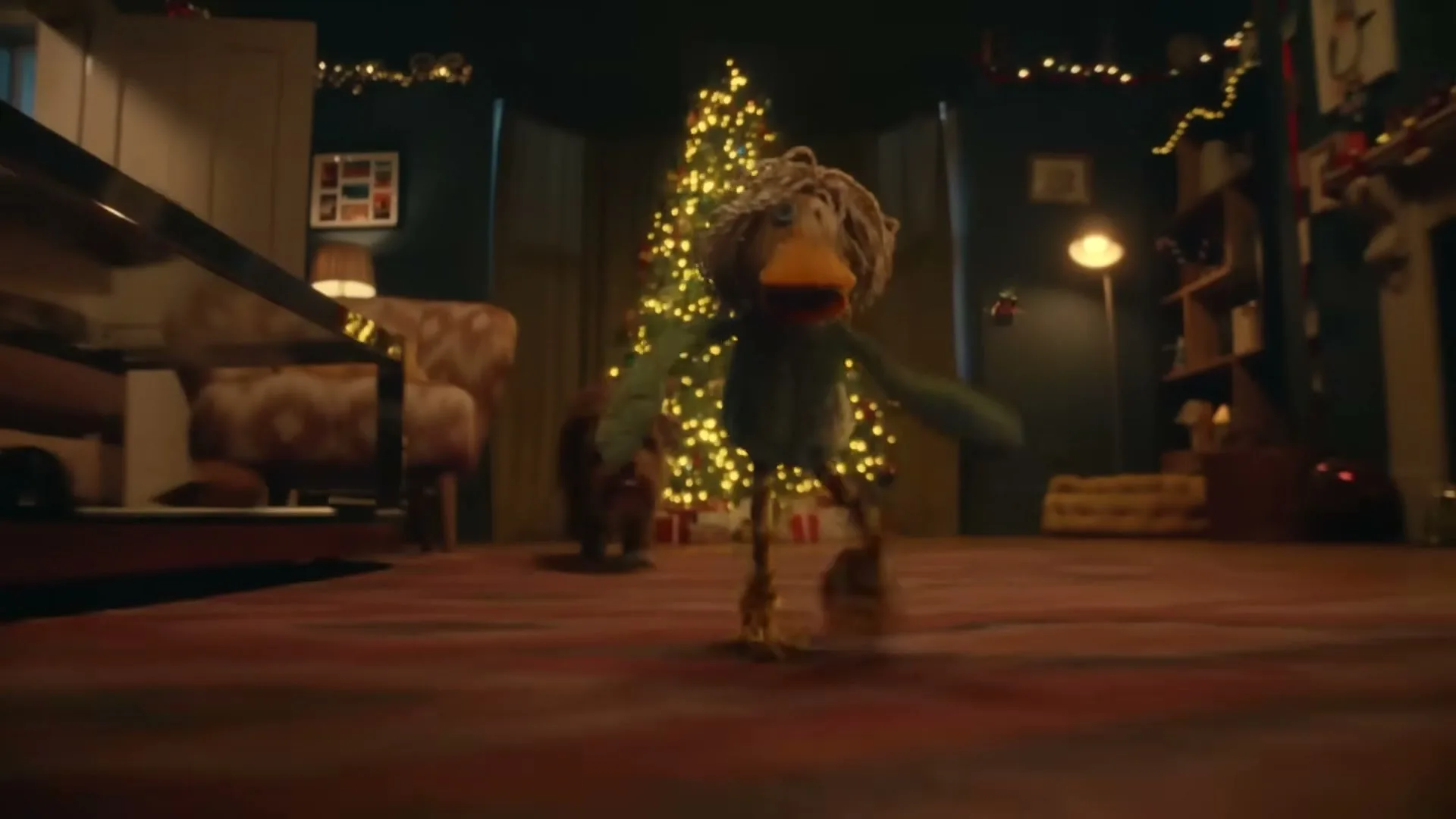 A stop-motion animated duck with a mop-like head and green body runs towards the camera in a festive living room with a Christmas tree.