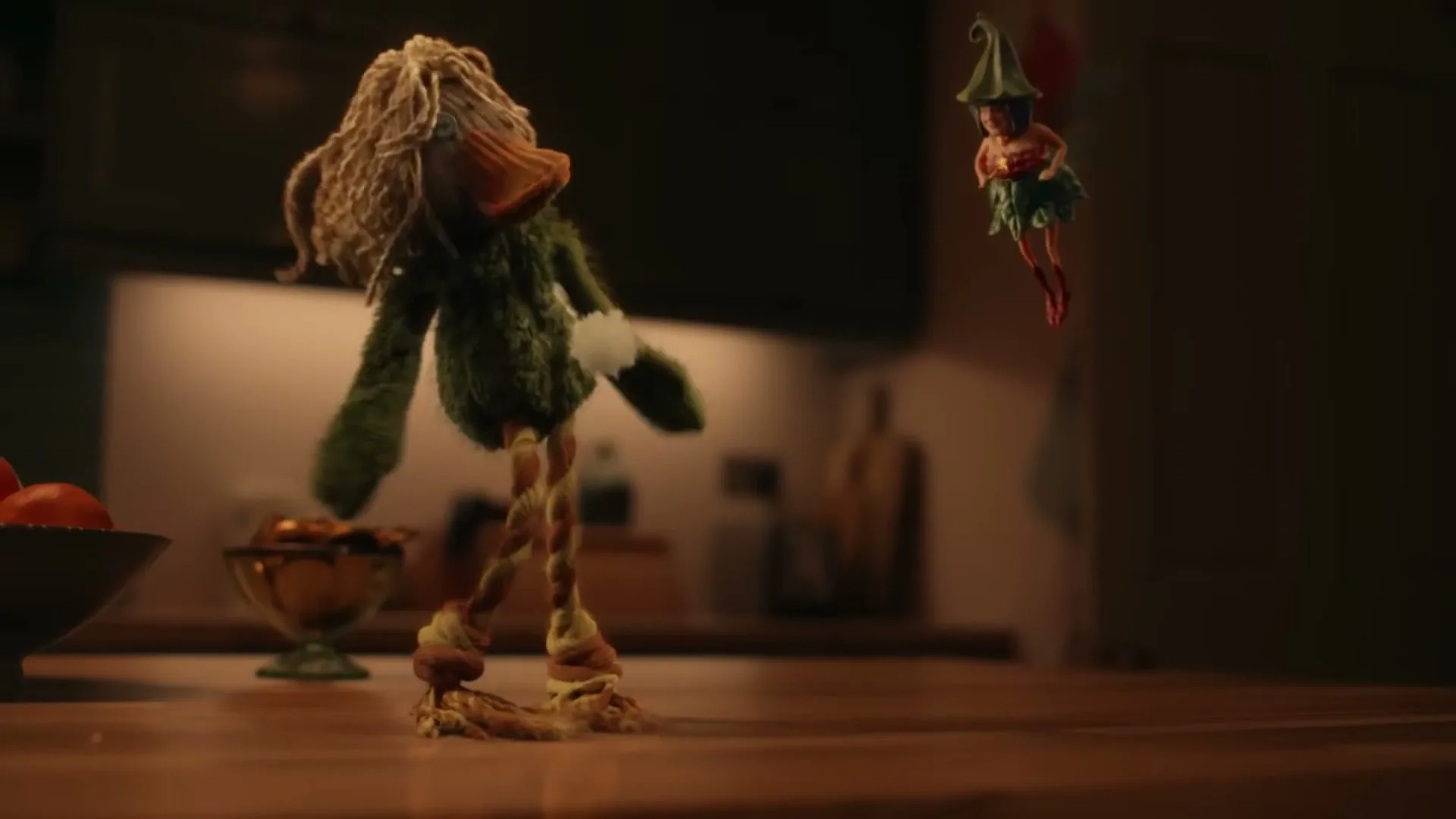 A green duck-like puppet with blonde hair falls onto a kitchen counter, losing a piece of fluff, while a small fairy puppet floats nearby.