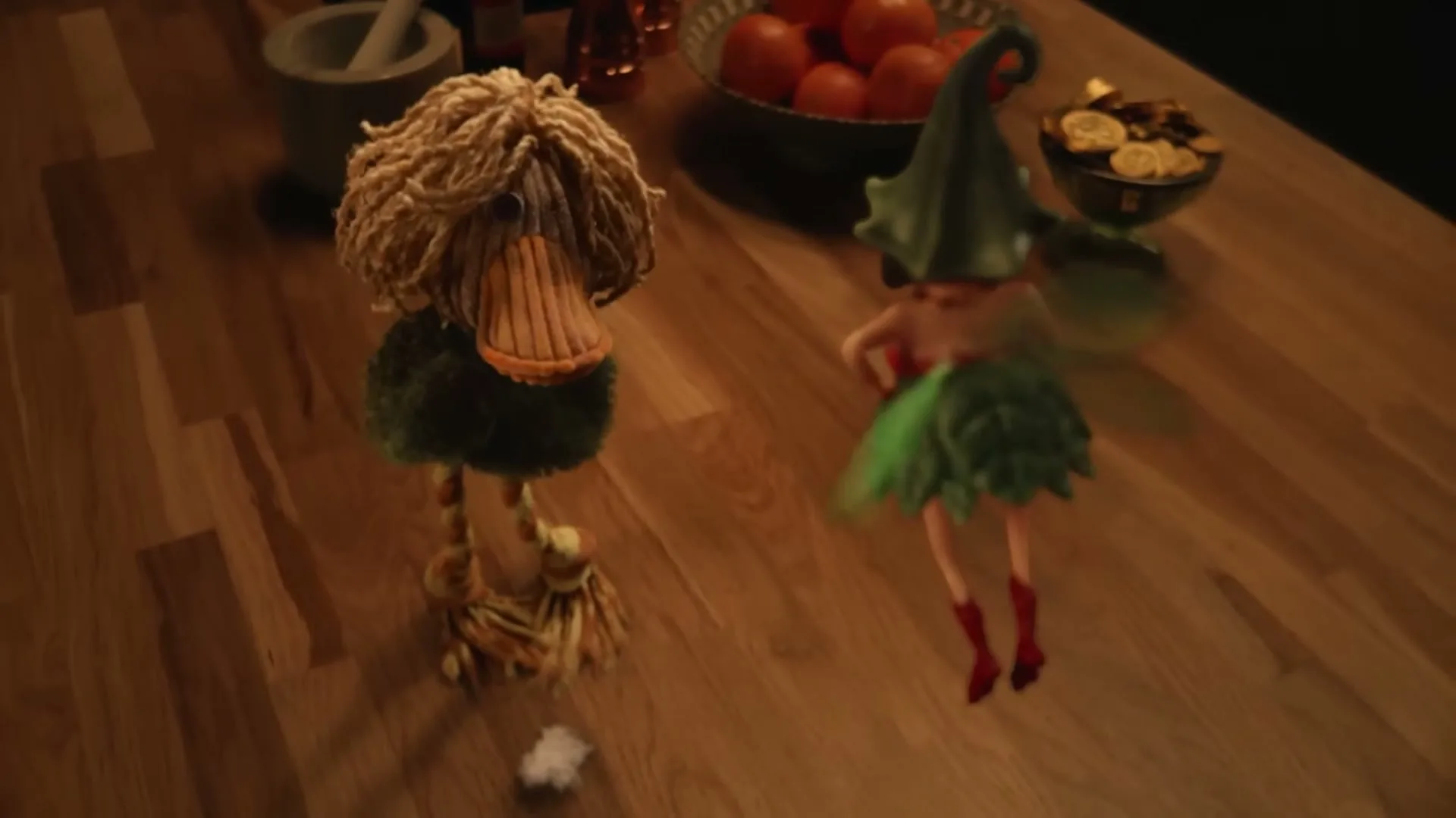 A stop-motion animated duck-like creature with shaggy hair and a fairy with green attire stand on a wooden table, looking at each other.