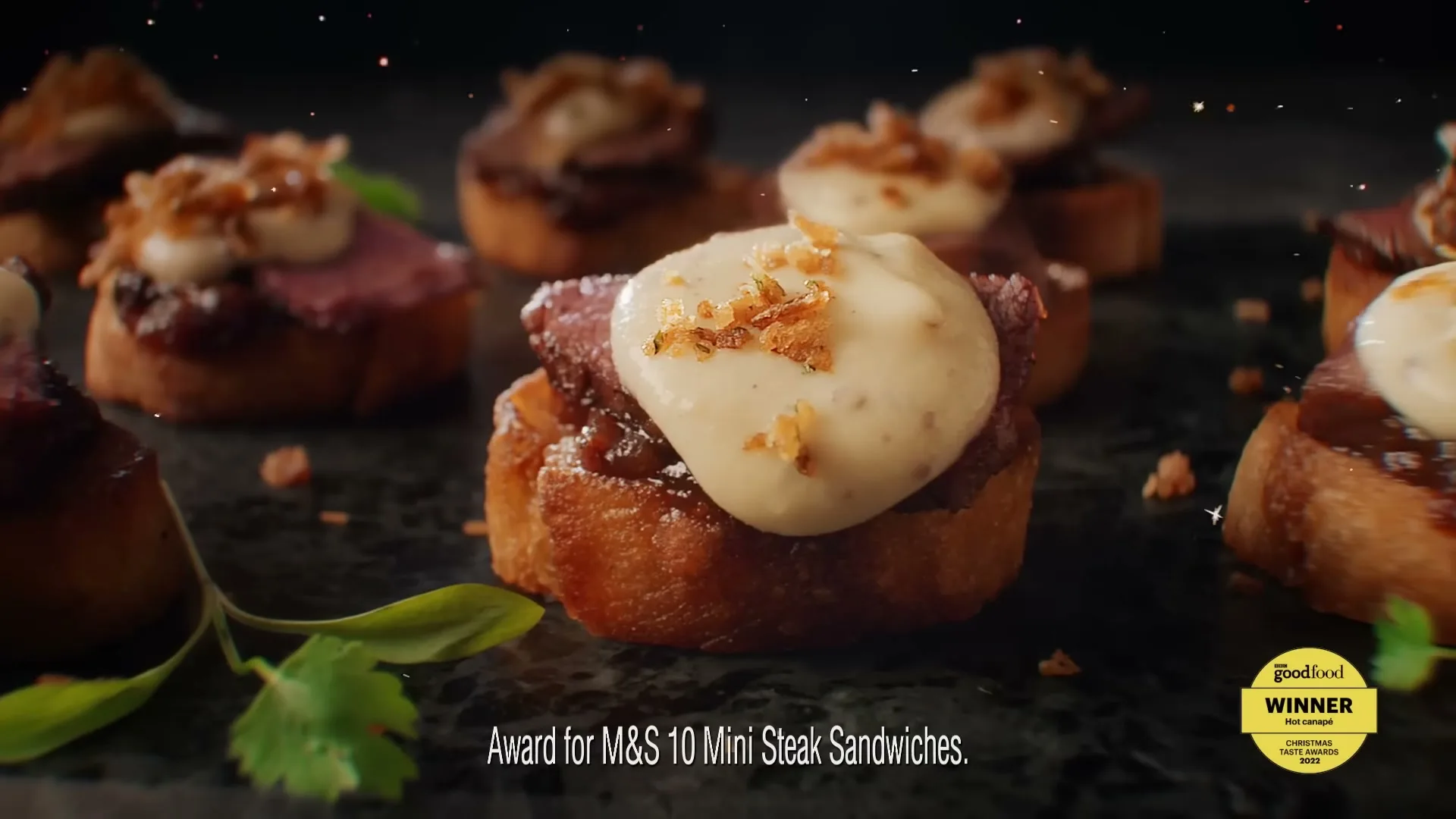 A close-up shot of several mini steak crostinis with caramelized onions and a creamy topping, garnished with fresh herbs, on a dark surface.