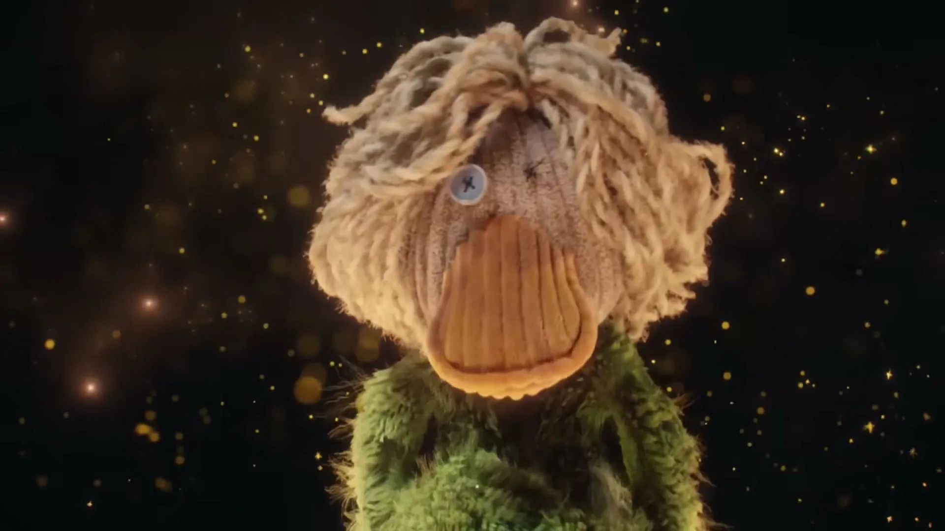 A green duck puppet with a button eye and blonde yarn hair looks around, surrounded by golden sparkles on a dark background.