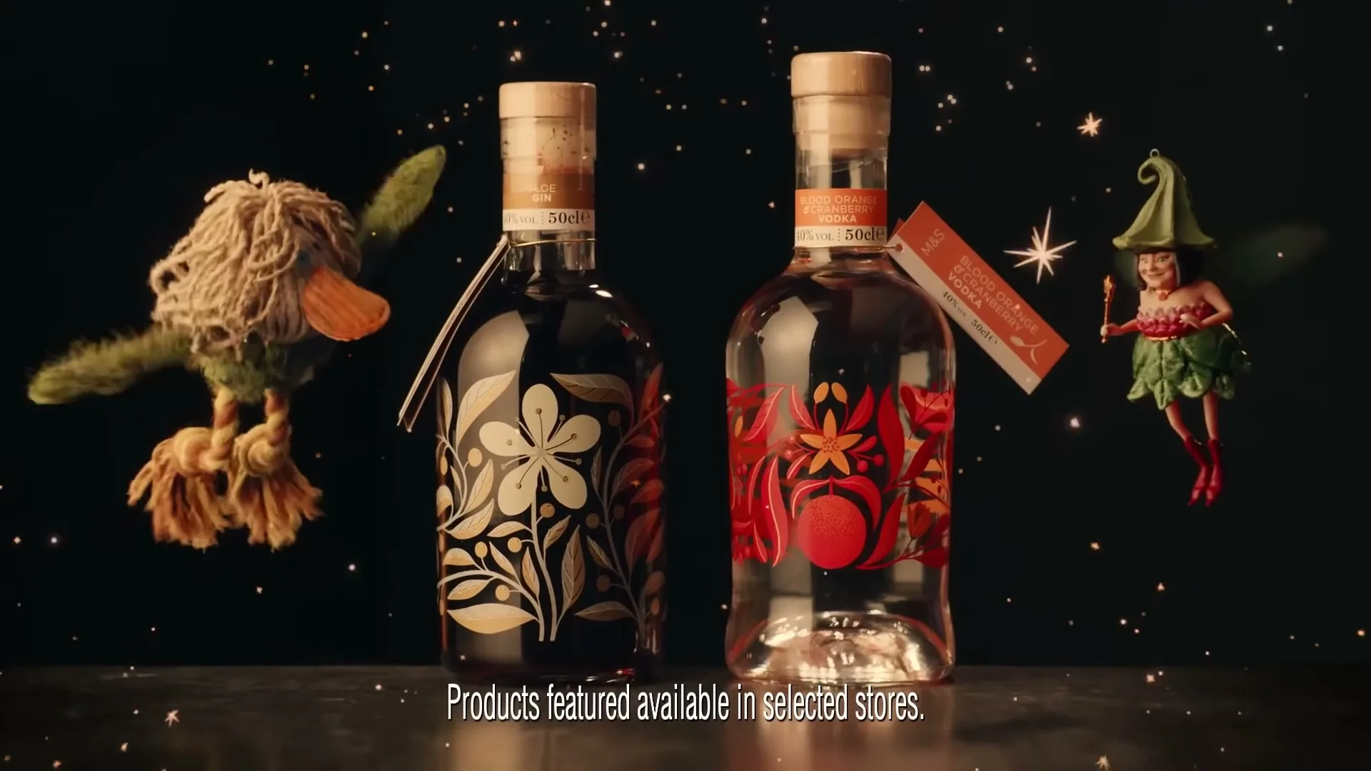 Two M&S gin bottles with decorative labels stand on a dark surface, flanked by a flying duck puppet and a fairy puppet, against a black background with sparkling lights.