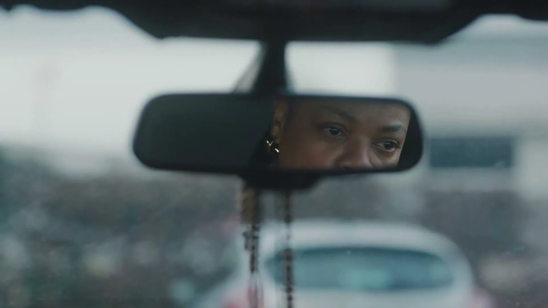 A woman with short hair and an earring is seen in a car's rearview mirror, looking down with a serious expression on a cloudy day.