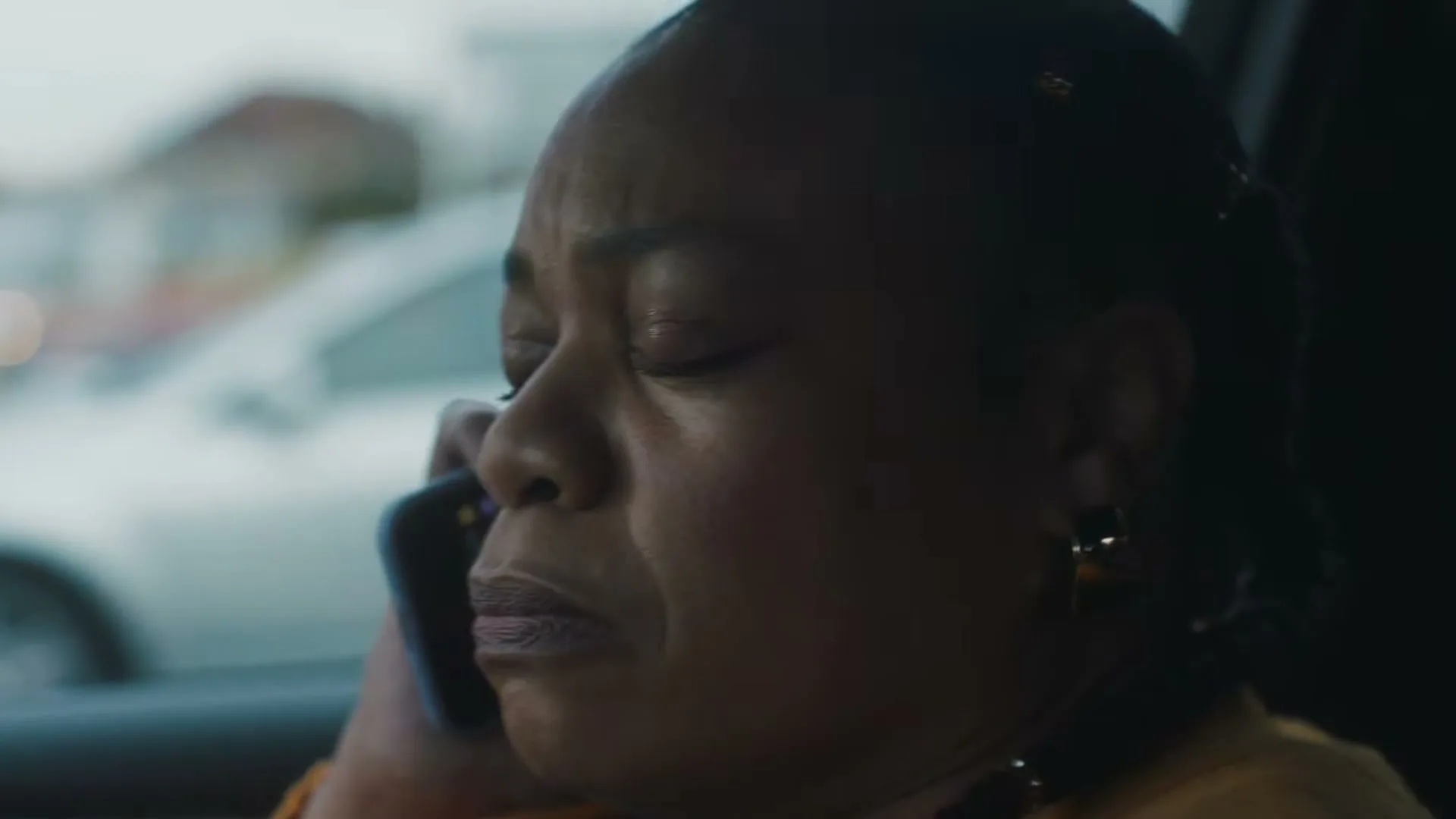 A woman in her 50s, wearing an orange cardigan, is seen in a medium closeup, talking on a phone in a car, looking sad.