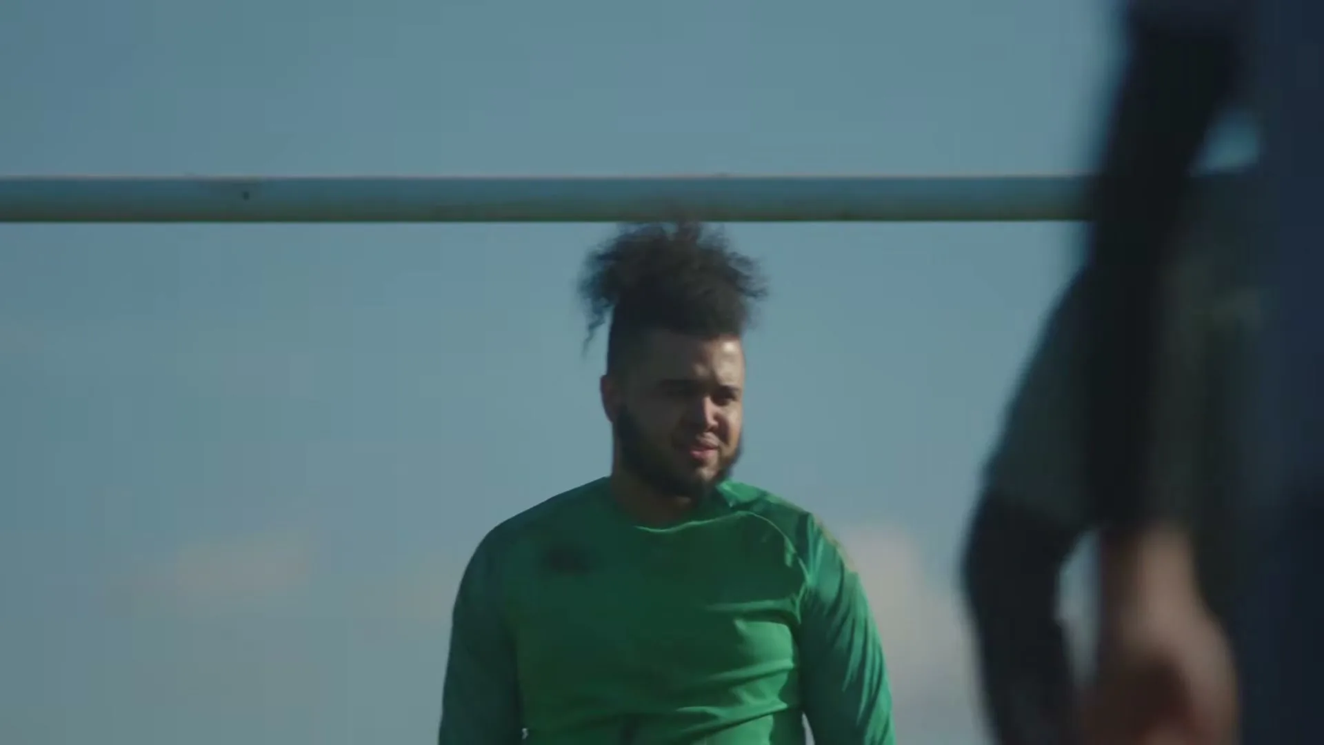 A young man with curly hair and a beard, wearing a green shirt, looks around on a sunny day at an outdoor sports field.